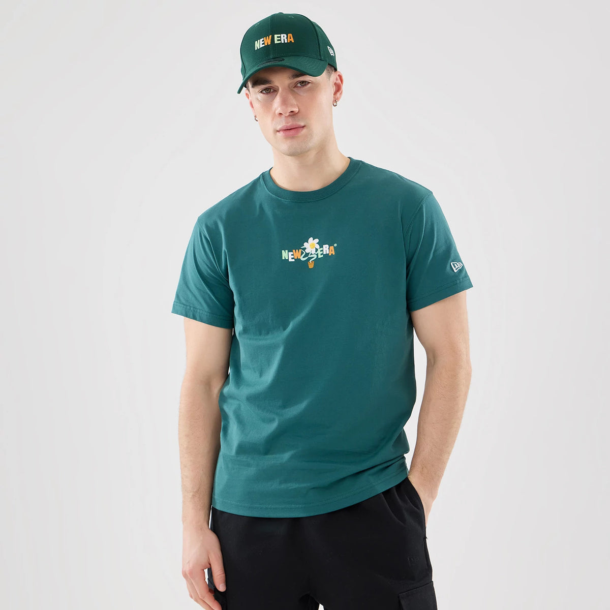New Era Earth Day Dark Green T-Shirt, on-model view showing a dark green shirt with a colourful New Era logo centred on the chest and a small raised New Era logo on the left sleeve, paired with a matching dark green cap.