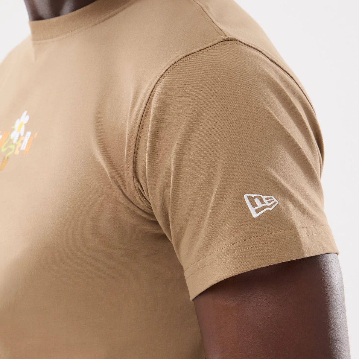 New Era Earth Day Beige T-Shirt, side view showing the short sleeve with a white embroidered New Era logo near the cuff and part of a floral graphic on the chest against the soft beige fabric.