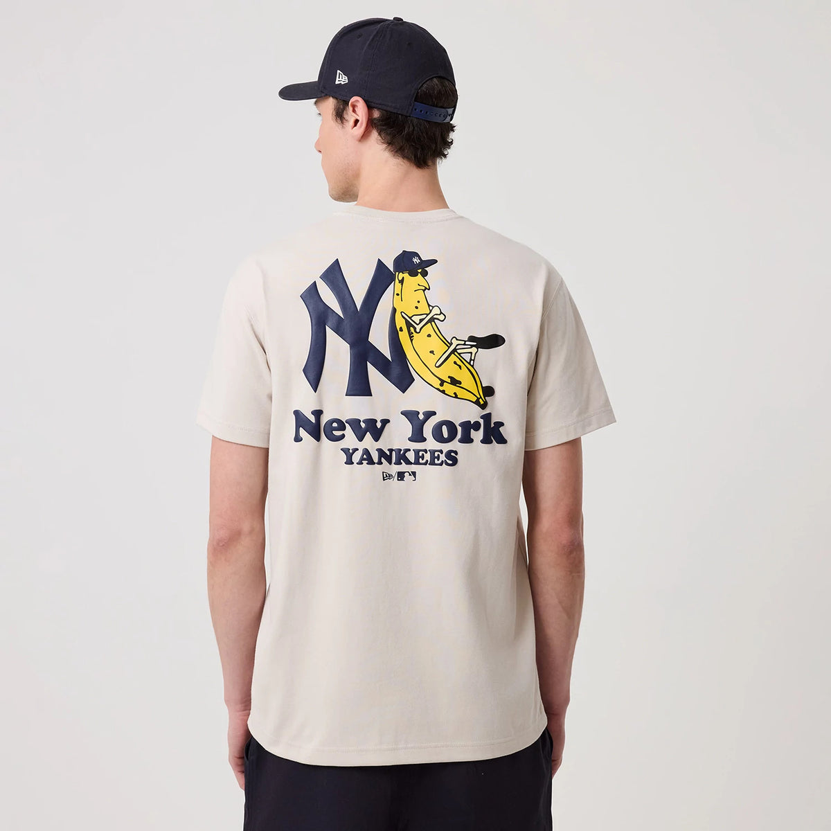 New York Yankees MLB Banana Graphic Natural T-Shirt, rear view showing a large navy New York Yankees logo with a yellow banana graphic leaning against it, navy text reading "New York Yankees" printed below on a natural coloured background.
