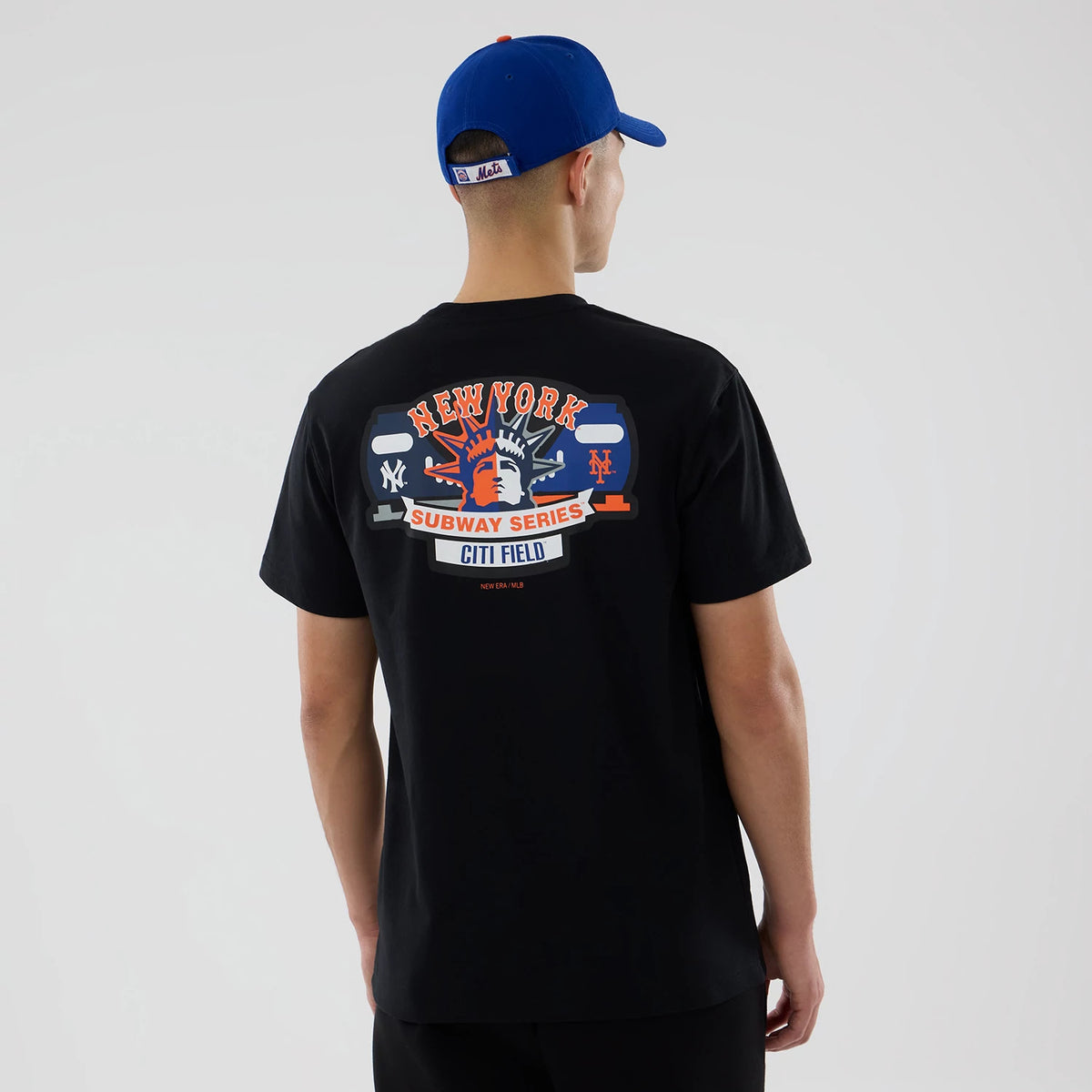 New York Mets MLB City Series Black T-Shirt, rear view showing a colourful graphic print featuring the Statue of Liberty, New York Yankees, and Mets logos on a black background, worn by a person in a blue cap.