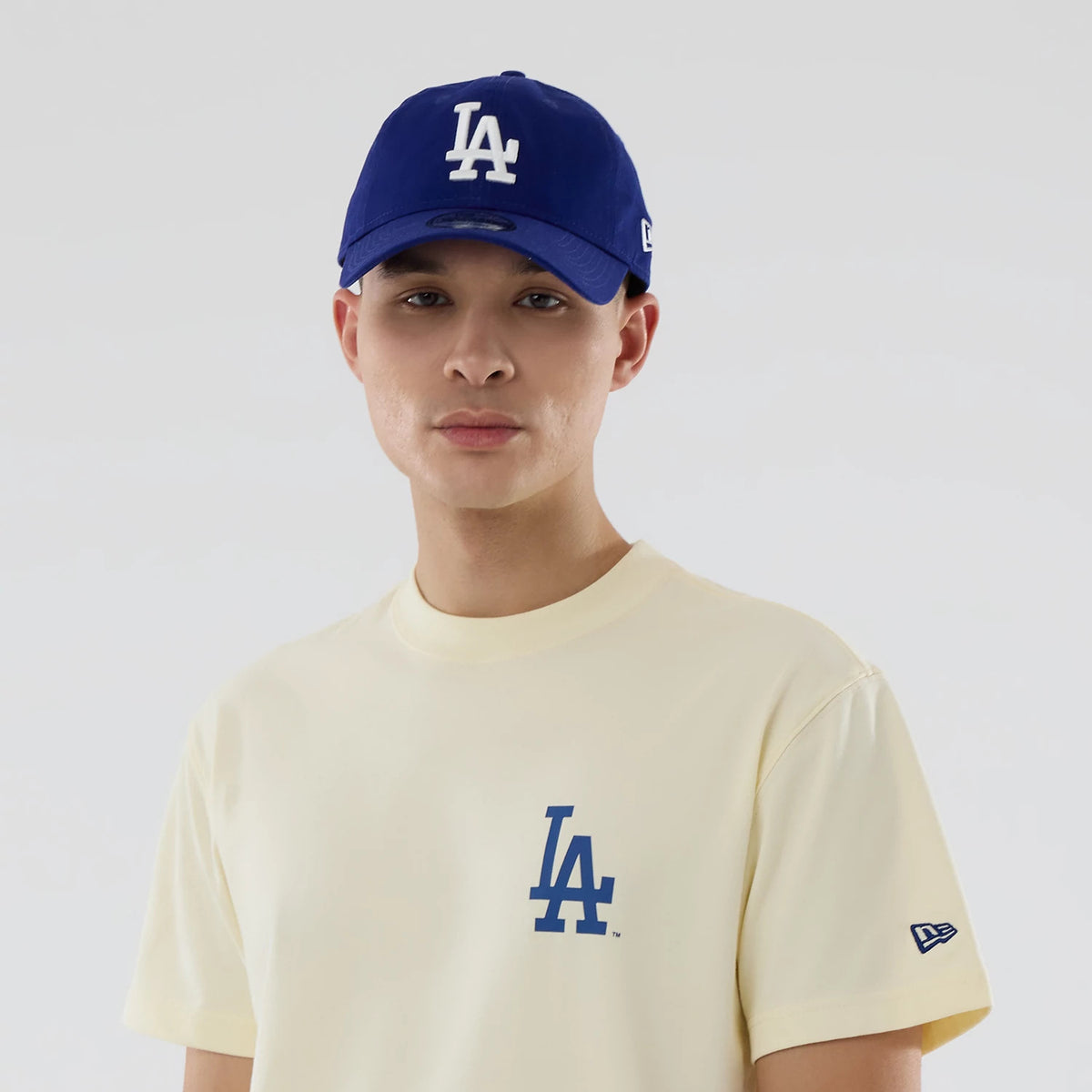 LA Dodgers MLB City Series Cream T-Shirt, on-model view showing the cream shirt with a blue LA logo on the chest, worn with a blue cap featuring a raised white LA logo, and a New Era logo on the sleeve.