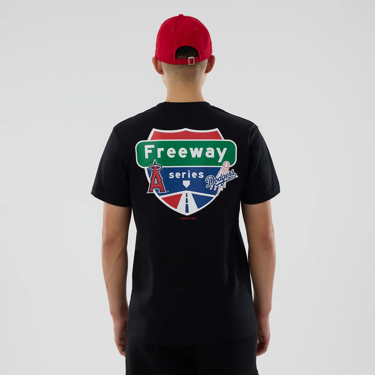 LA Angels MLB City Series Black T-Shirt, rear view showing a large freeway sign design with team logos across the back, worn by a person in a red cap, the black fabric providing a bold contrast to the colourful graphic.