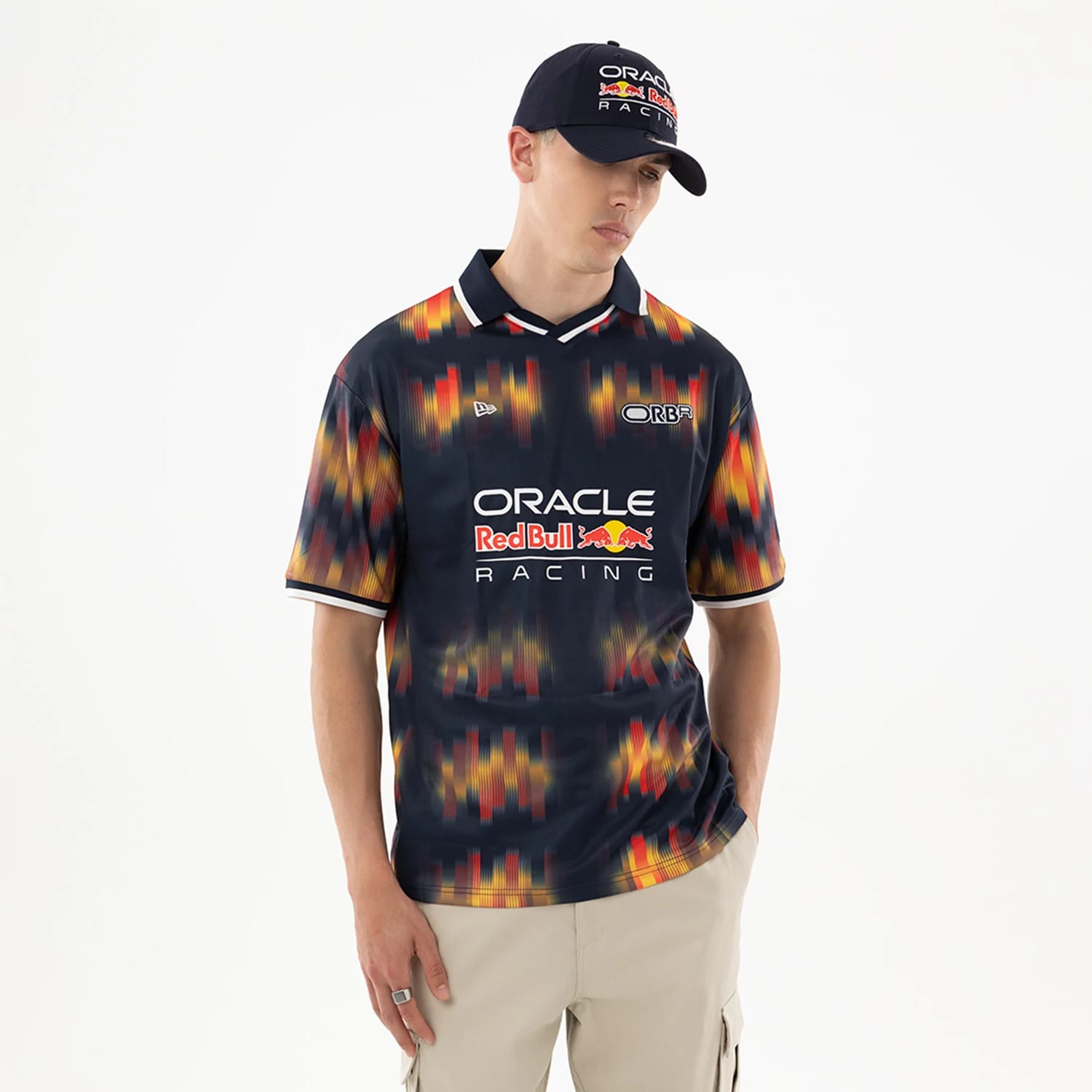 Red Bull Racing Multi Soccer Jersey, front view on a model wearing a matching cap, displaying the Oracle Red Bull Racing logo prominently on the chest with dynamic multicoloured patterning on the sleeves and body.