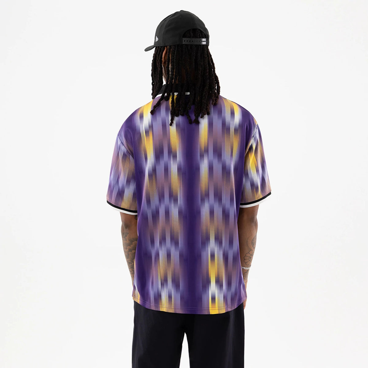 LA Lakers NBA Purple Soccer Jersey, back view on a model wearing a dark cap with a New Era flag logo, displaying a bold purple and yellow pattern with black sleeve trim and a black collar.