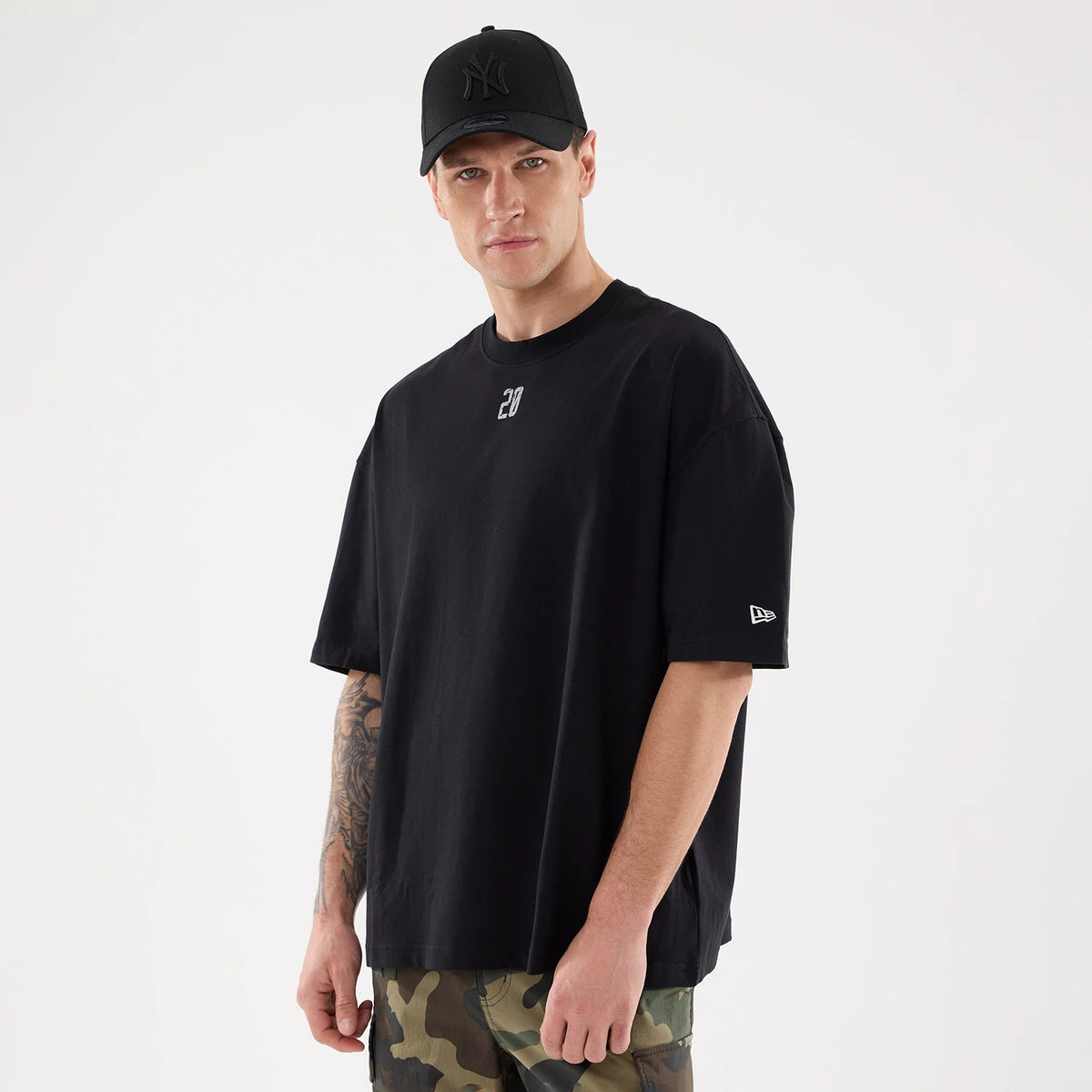 New Era Graphic Street Archived City Black Oversized T-Shirt, on-model view featuring a loose black t-shirt with a small white '20' on the chest and a white embroidered New Era logo on the left sleeve, paired with a black cap.