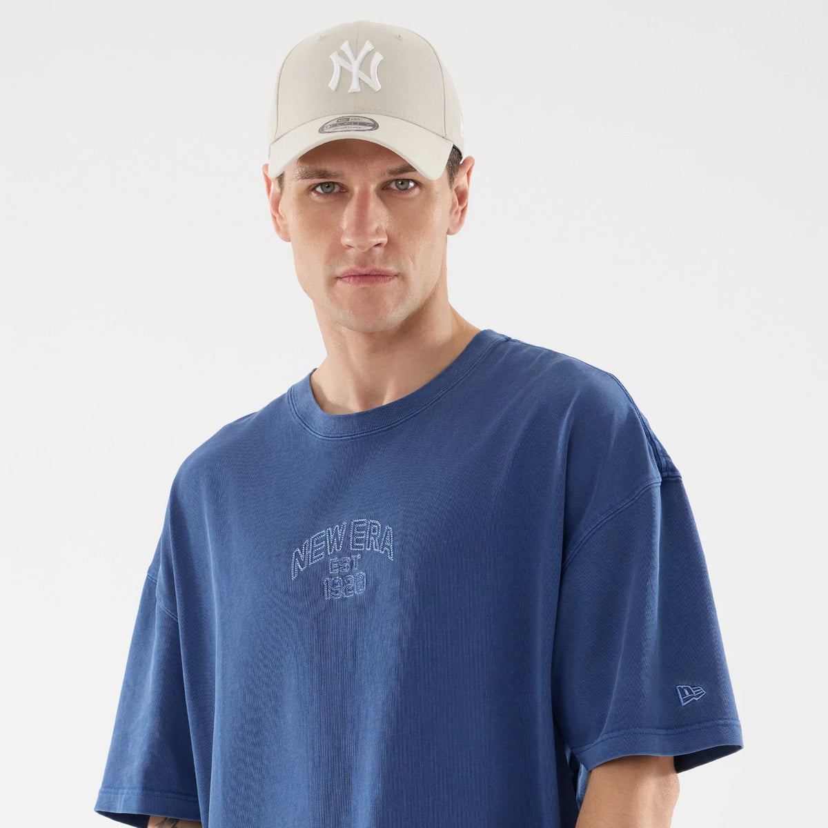 New Era Graphic Street Archived Blue Oversized T-Shirt, on-model view showing the embossed New Era EST 1920 logo on the chest and a small raised New Era logo on the left sleeve, worn with a cream cap featuring a raised white NY logo.