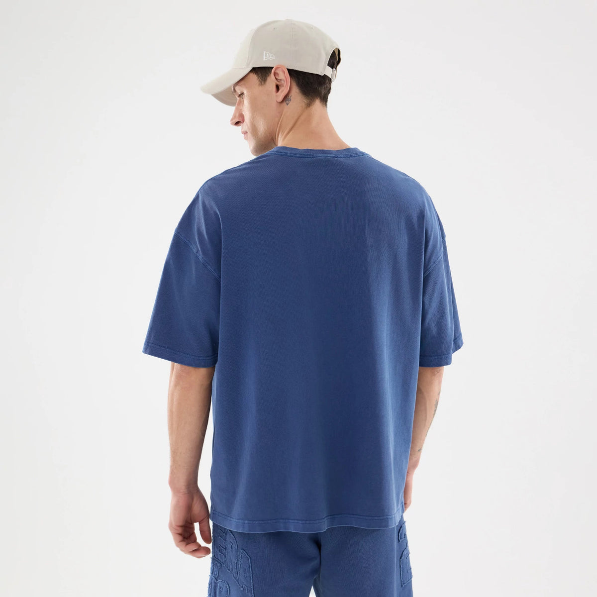 New Era Graphic Street Archived Blue Oversized T-Shirt, rear view showing the loose fit of the blue fabric with short sleeves, worn by a model in matching blue shorts and a beige cap with an embossed logo on the side.
