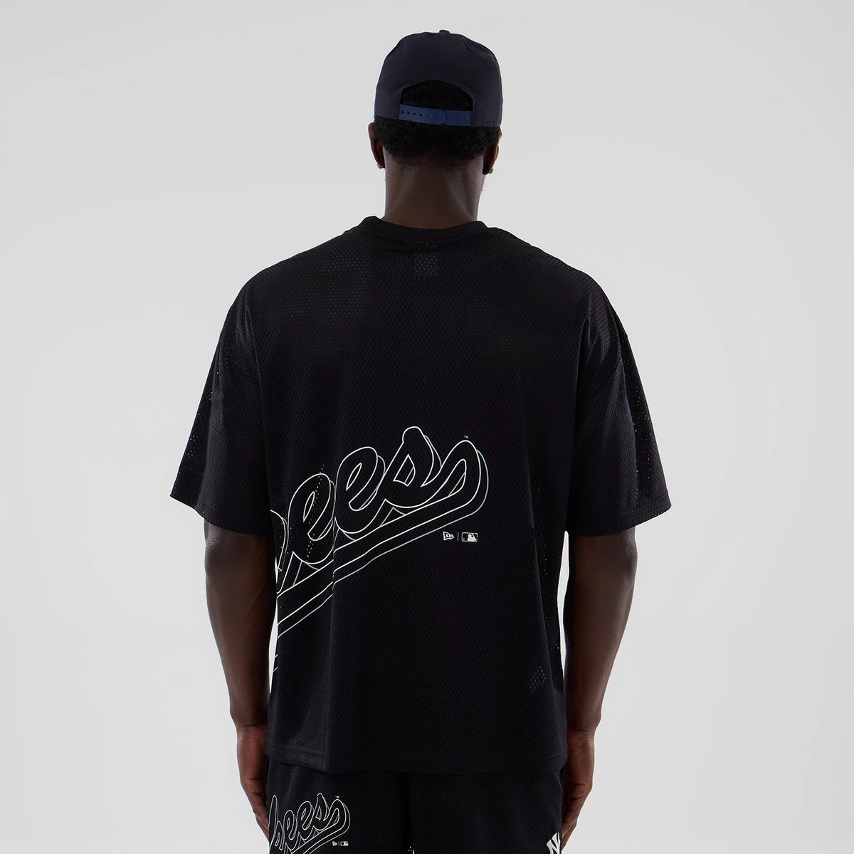 New York Yankees MLB Mix League Black Oversized Mesh T-Shirt, rear view showing a large Yankees script across the back in white, worn by a model in a black cap, and featuring small embroidered logos near the lower hem.