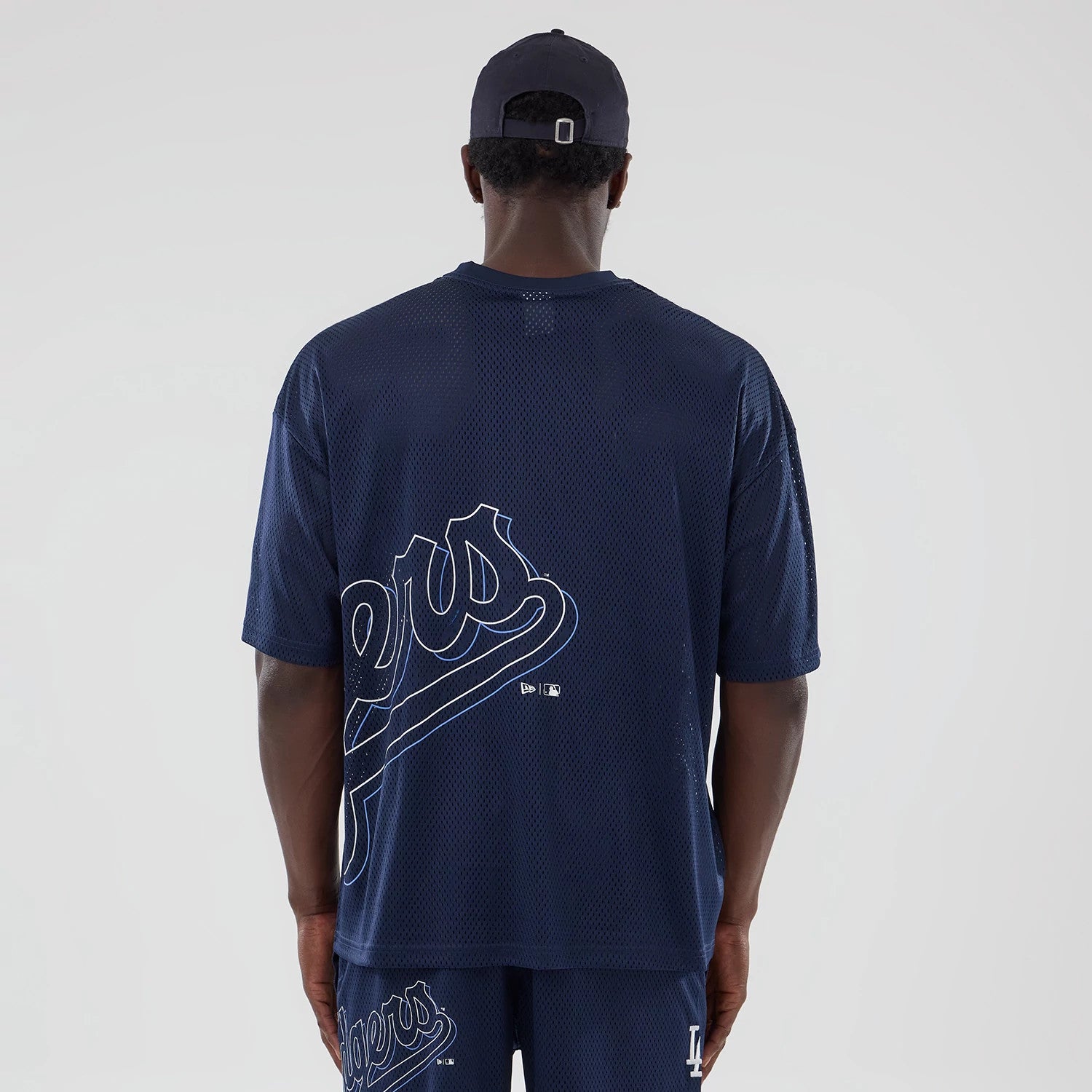 LA Dodgers MLB Mix League Blue Oversized Mesh T-Shirt, rear view showing large white Dodgers script across the back, small debossed New Era and MLB logos below the script, on-model with matching blue shorts and a black cap.