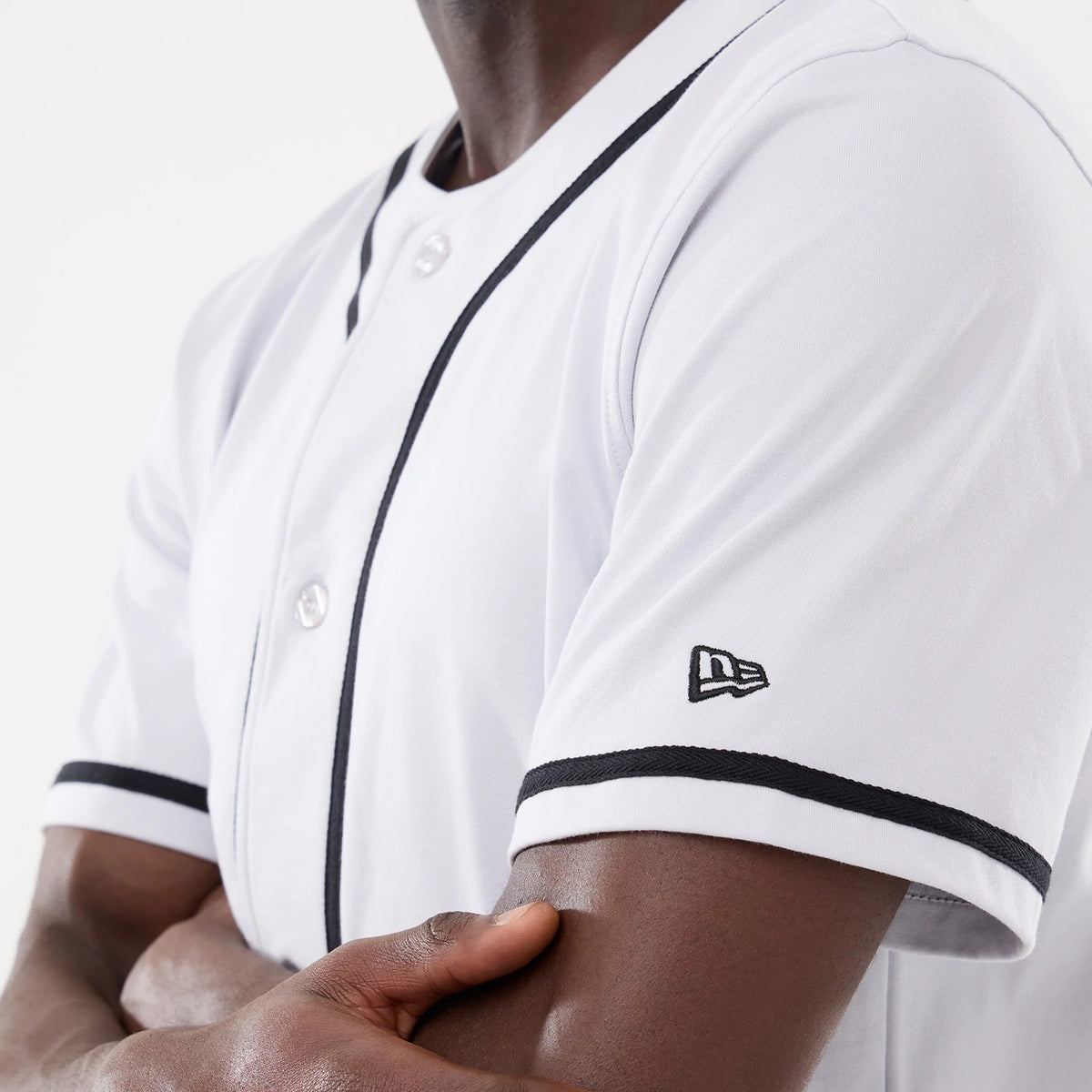New Era Basic White Jersey, side view on a model featuring black piping on the sleeves and down the front, with embroidered New Era flag logo on the left sleeve and white buttons down the centre.