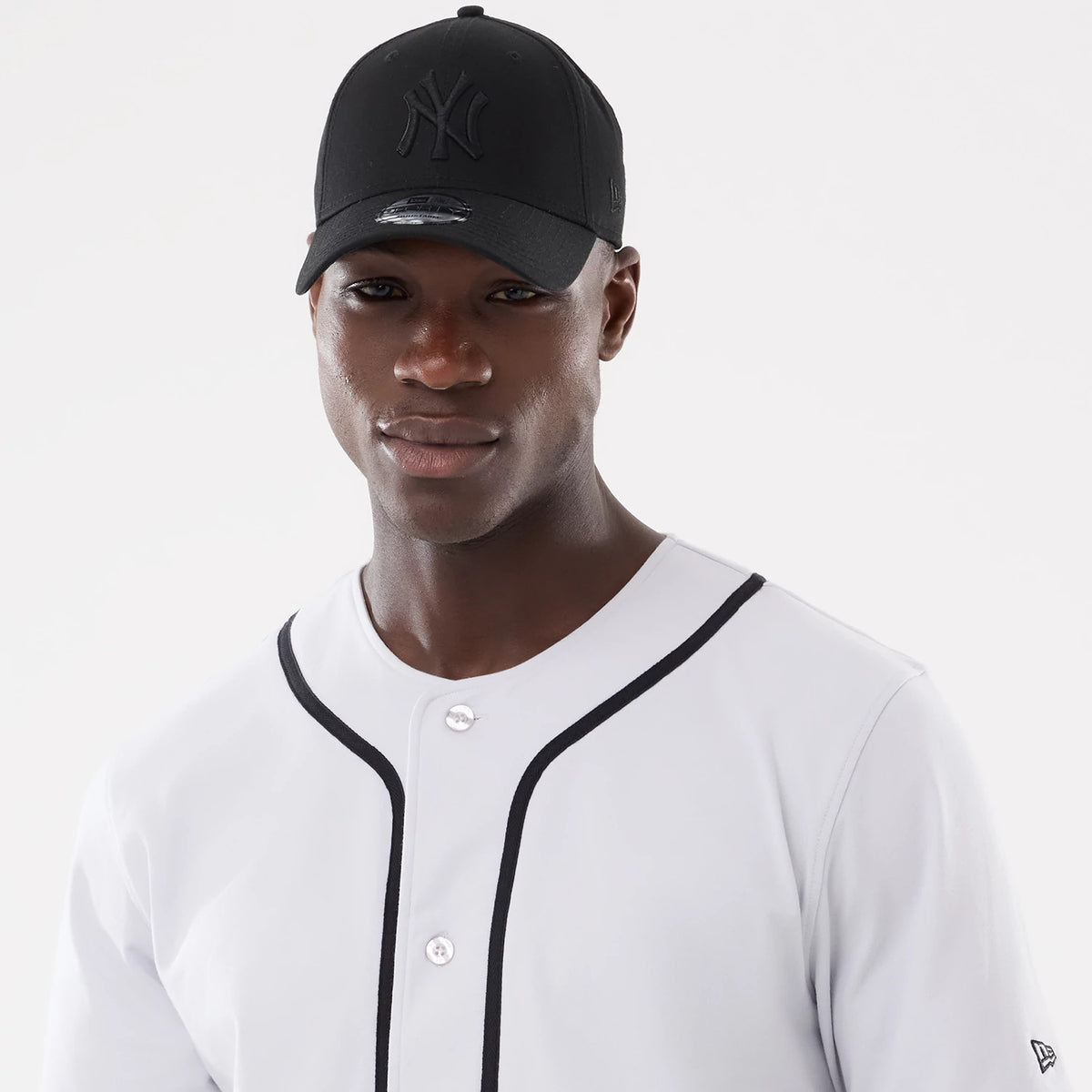 New Era Basic White Jersey, on-model view with a black cap featuring a raised embroidered NY logo on front and a curved visor with a 9FORTY size sticker, styled with a white jersey accented by black piping.