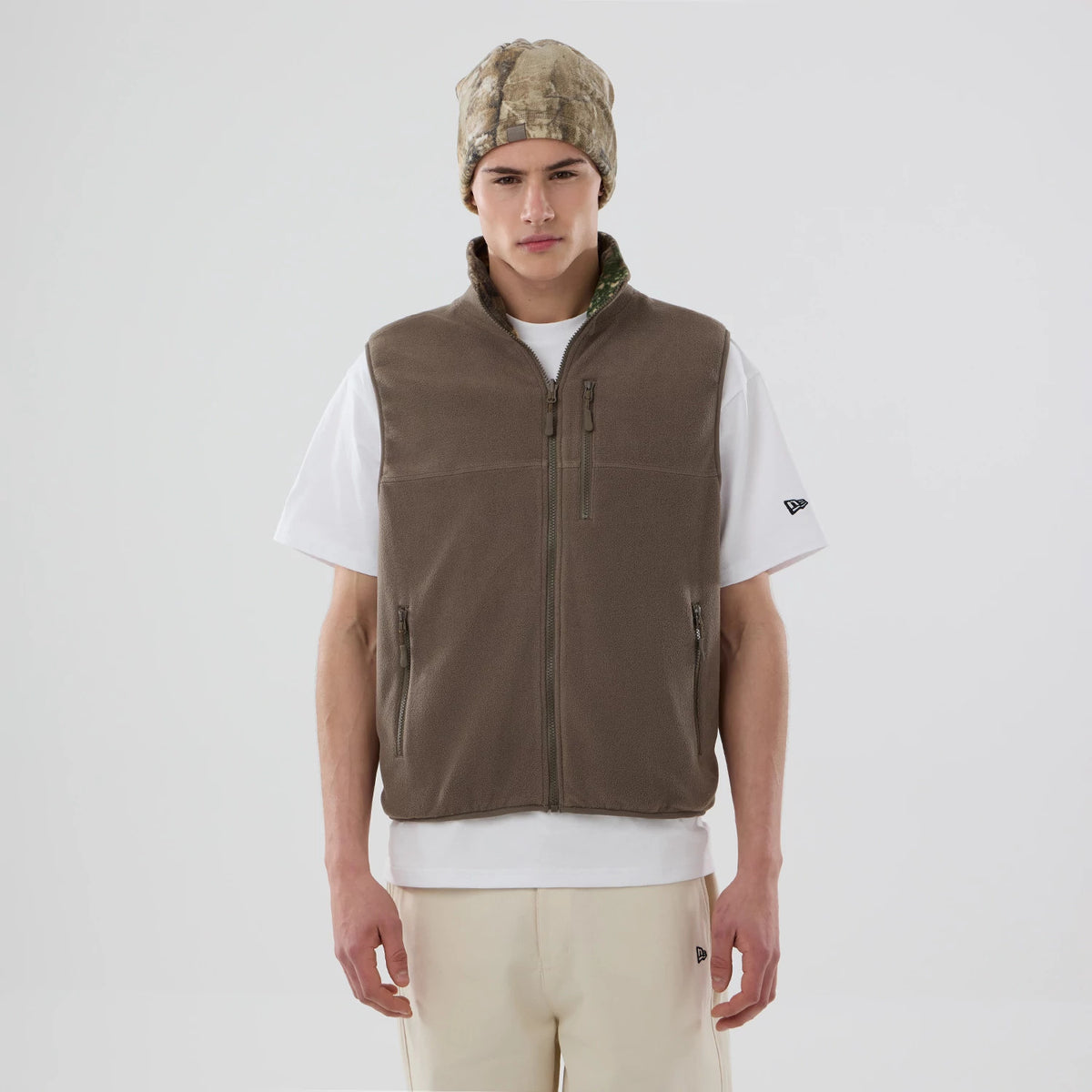 Thisisneverthat X New Era Revolution Fleece Real Tree Vest, front view on a model wearing a white tee, showing the structured zip-up design with side pockets and a zipped chest pocket, paired with a camouflage beanie.
