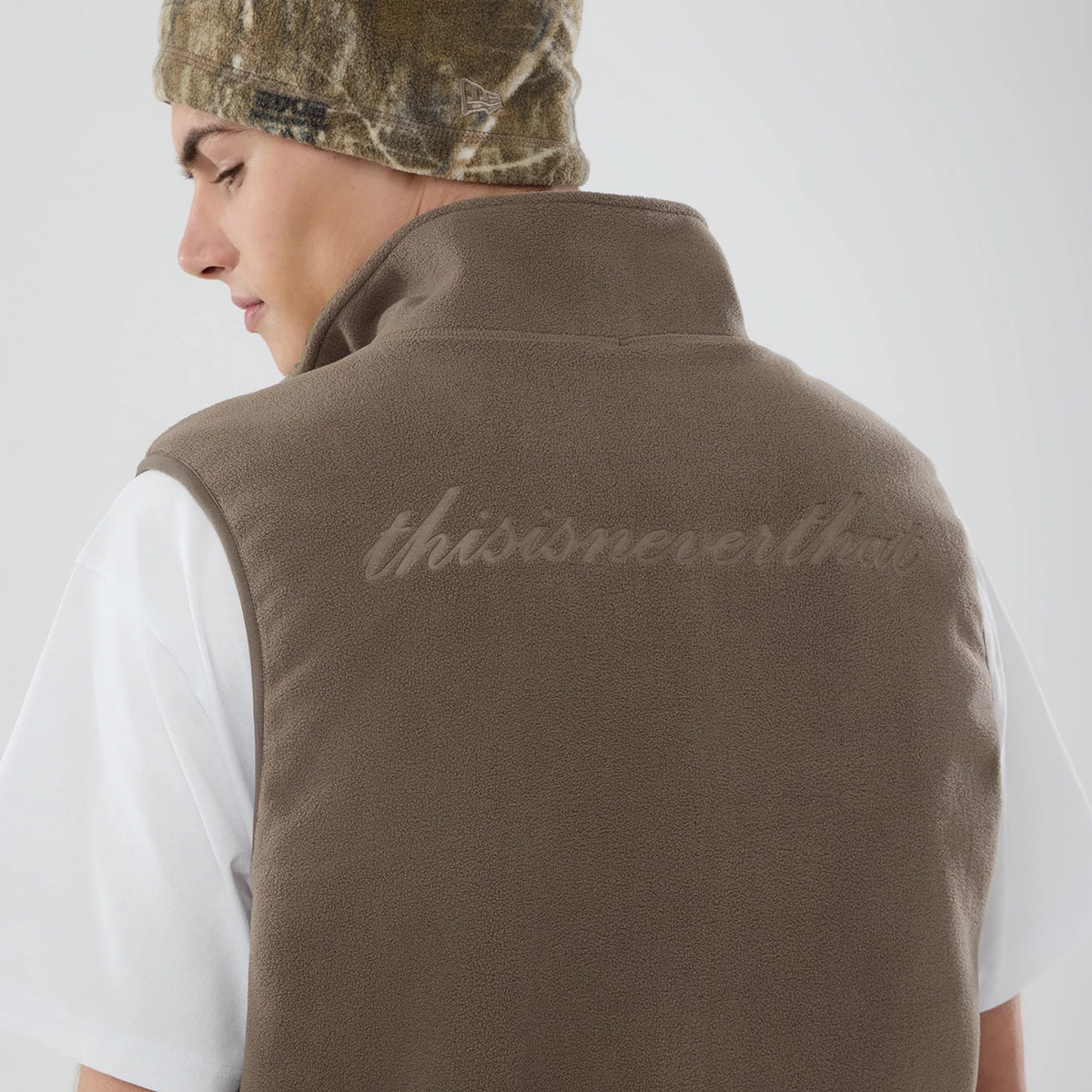 Thisisneverthat X New Era Revolution Fleece Real Tree Vest, rear view on a model wearing a camouflage beanie, featuring tonal thisisneverthat lettering across the upper back of the brown fleece fabric.