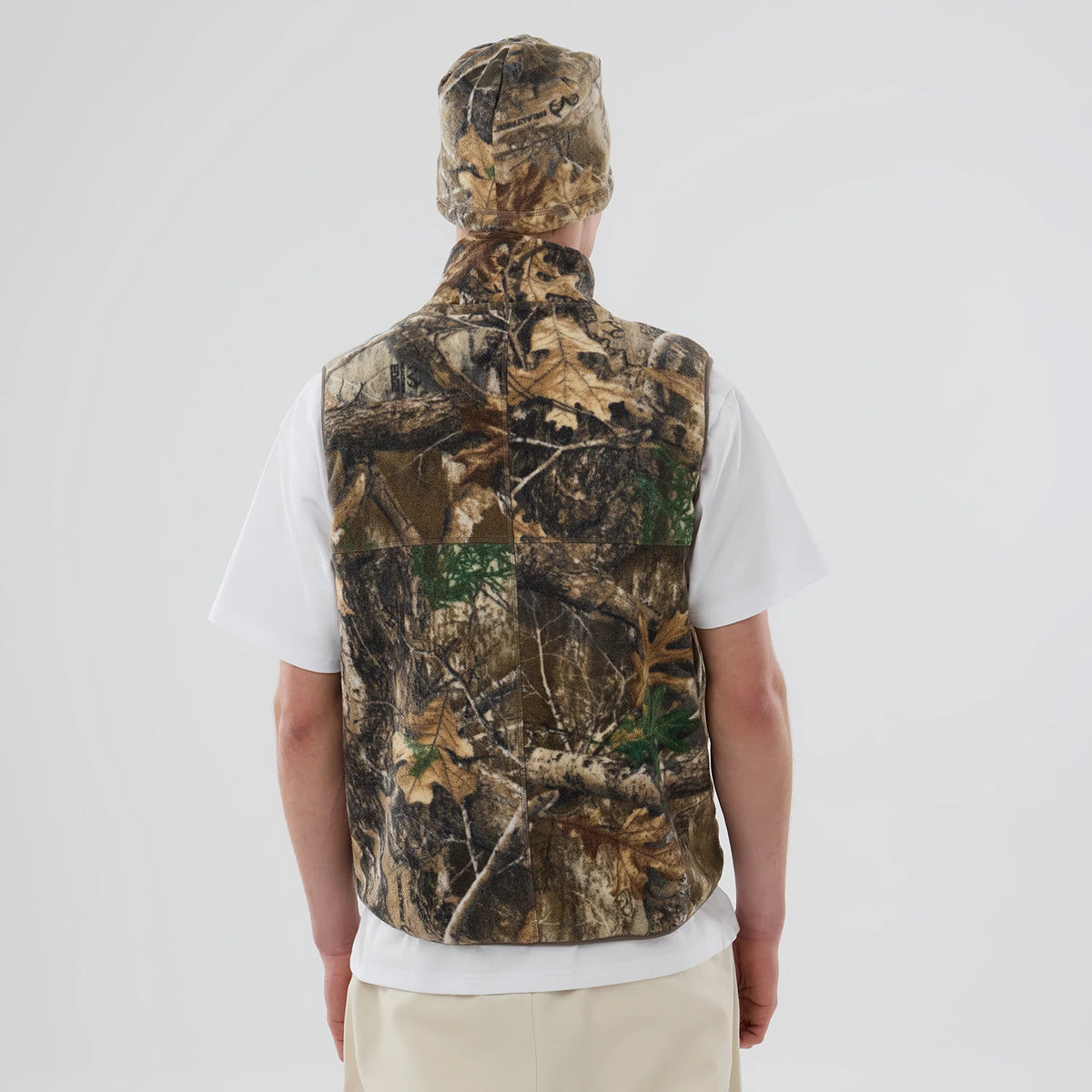 Thisisneverthat X New Era Revolution Fleece Real Tree Vest, rear view on a model wearing a matching camo hat and white T-shirt, displaying the vest's intricate woodland pattern and classic sleeveless cut.