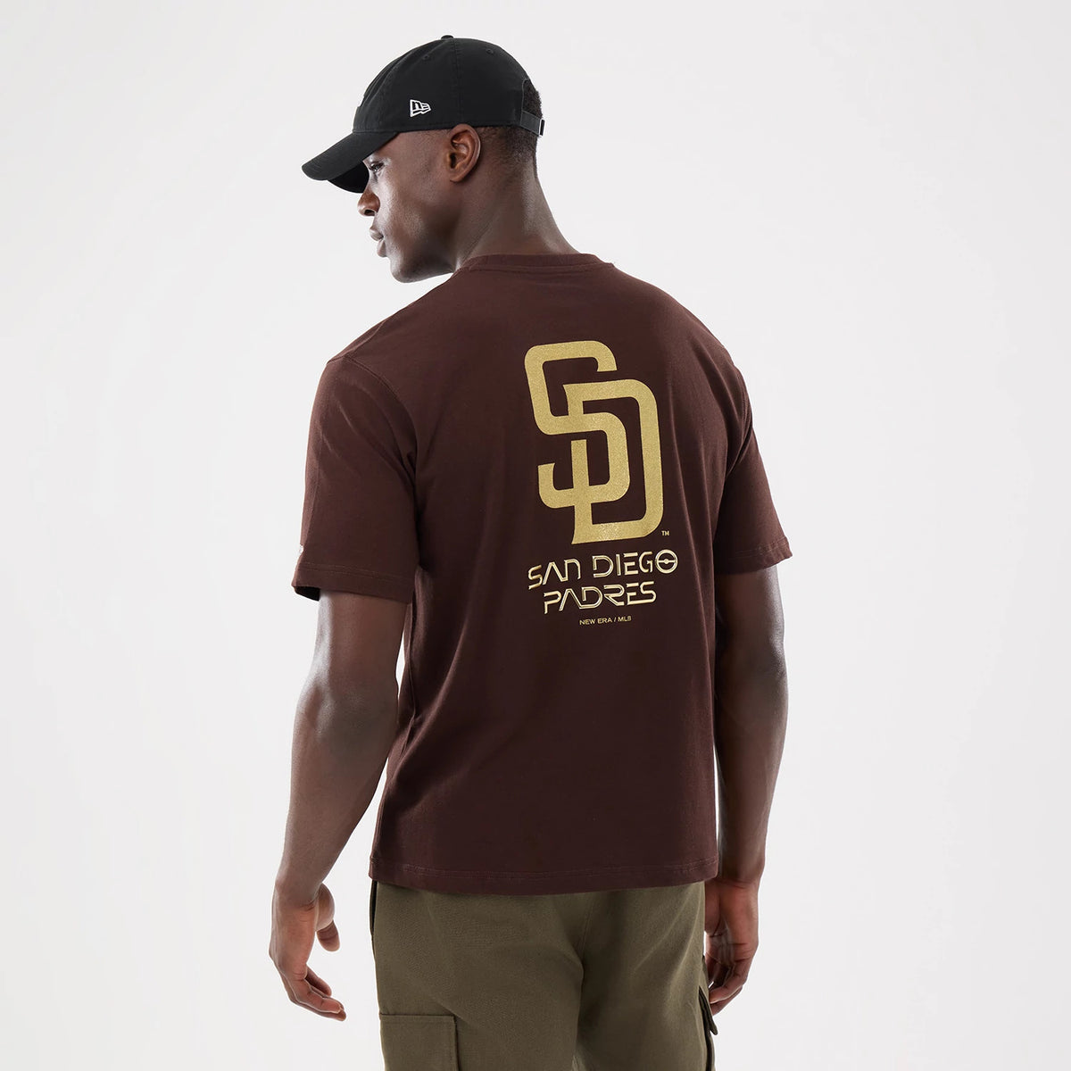 San Diego Padres Hajime Sorayama X MLB Dark Brown T-Shirt, rear view showing a gold "SD" logo centred on the back, with "San Diego Padres" text below, worn by a model in green trousers and a black cap with a raised logo on the side.