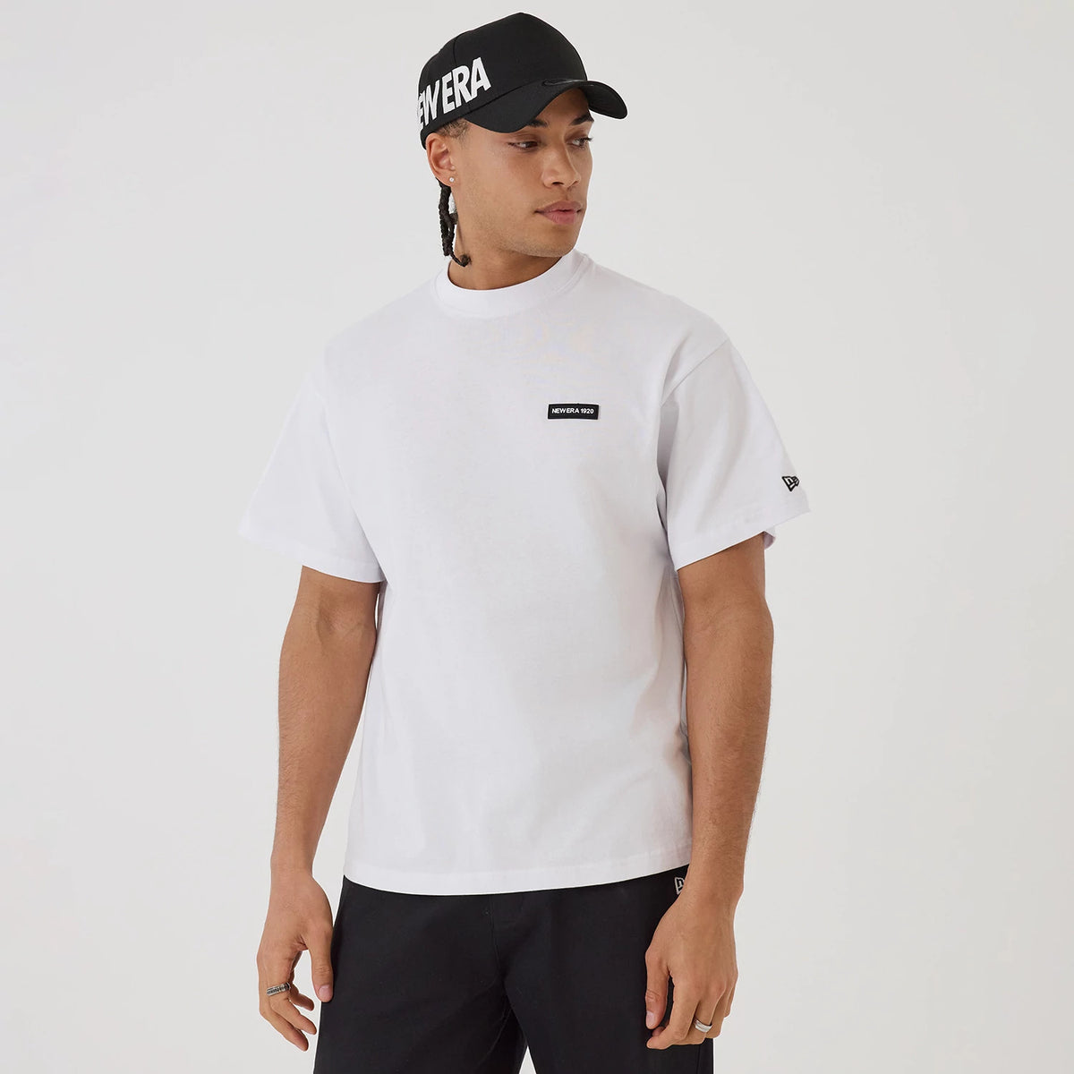 New Era Distressed White T-Shirt, on-model view featuring a clean white T-shirt with a black embossed New Era logo on the chest and a small logo on the left sleeve, paired with black trousers and a black cap displaying a raised New Era logo.