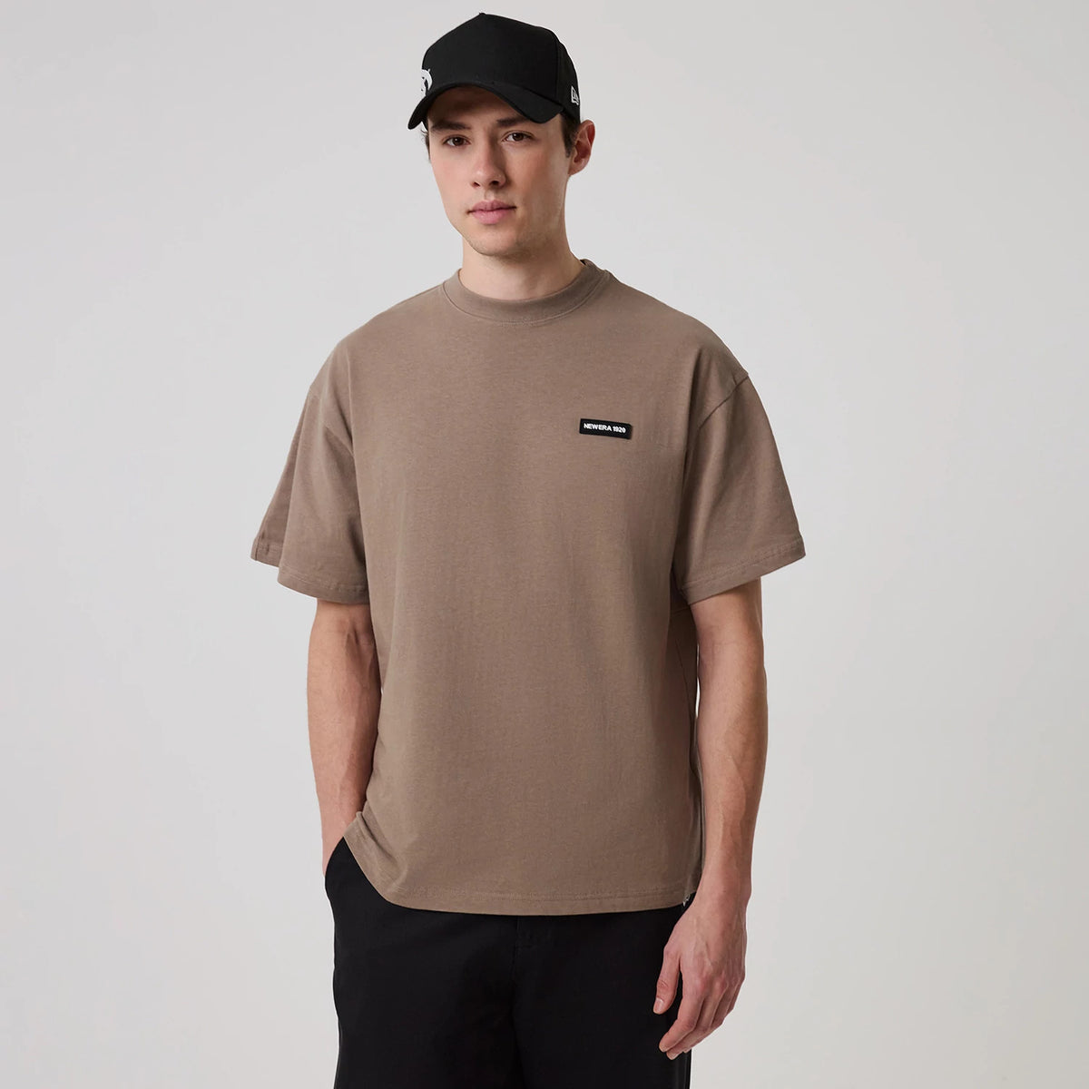 New Era Distressed Brown T-Shirt, on-model view showing a simple brown tee with short sleeves and a small black rectangular patch logo on the left chest, worn by a person also wearing a black cap.