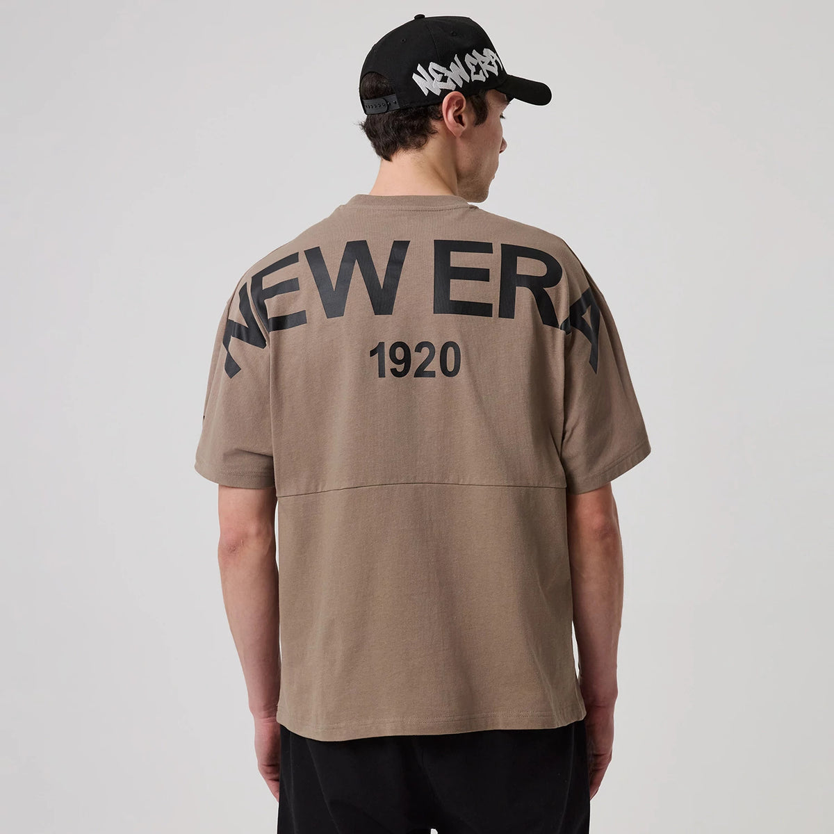 New Era Distressed Brown T-Shirt, rear view showing large black printed "NEW ERA 1920" text across the upper back, with the model wearing a black cap featuring a white raised logo positioned sideways.