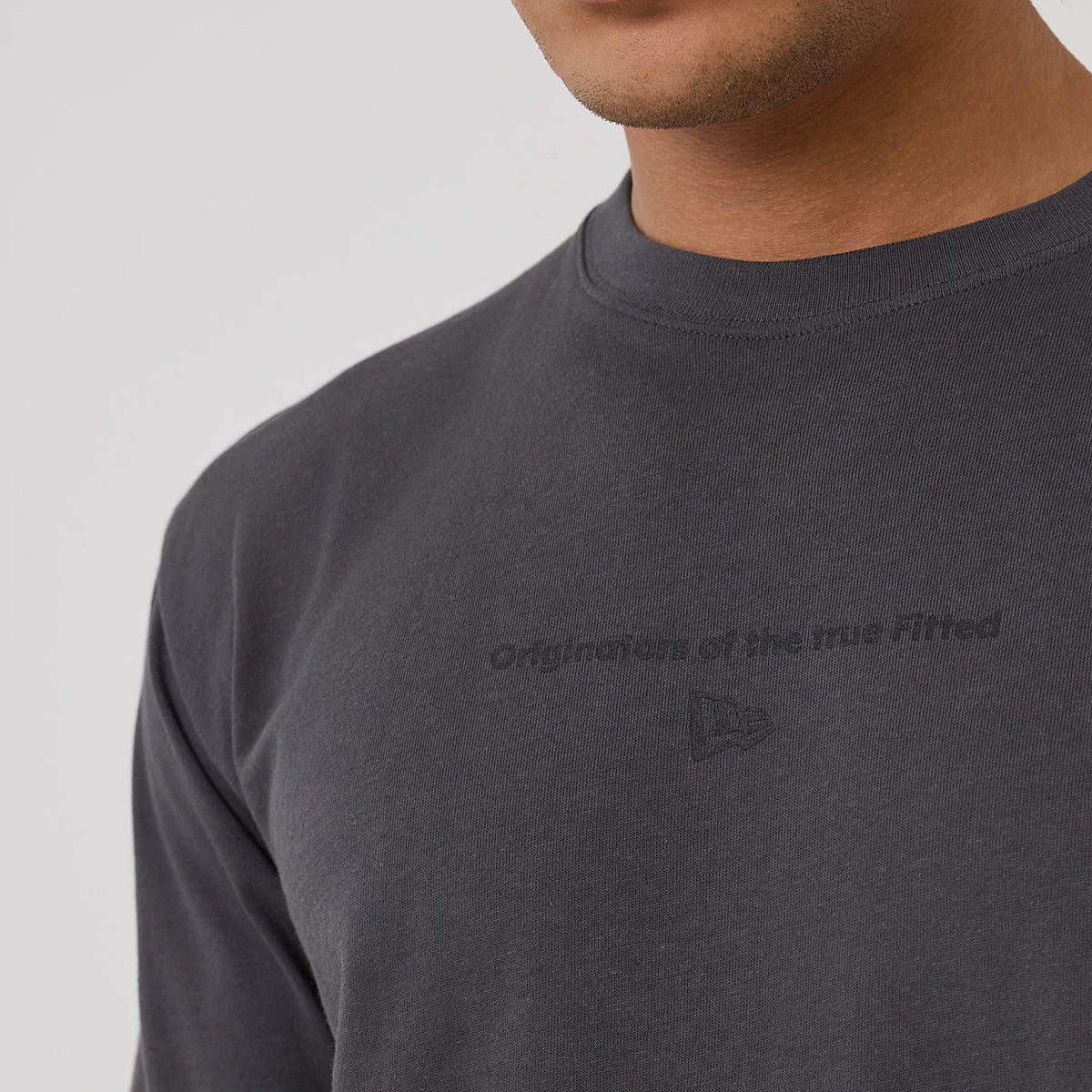 New Era Circle Grey T-Shirt, front view showing a dark grey fabric with a raised New Era logo and the wording 'Originators of the true fitted' embossed centrally on the chest near the crew neckline.