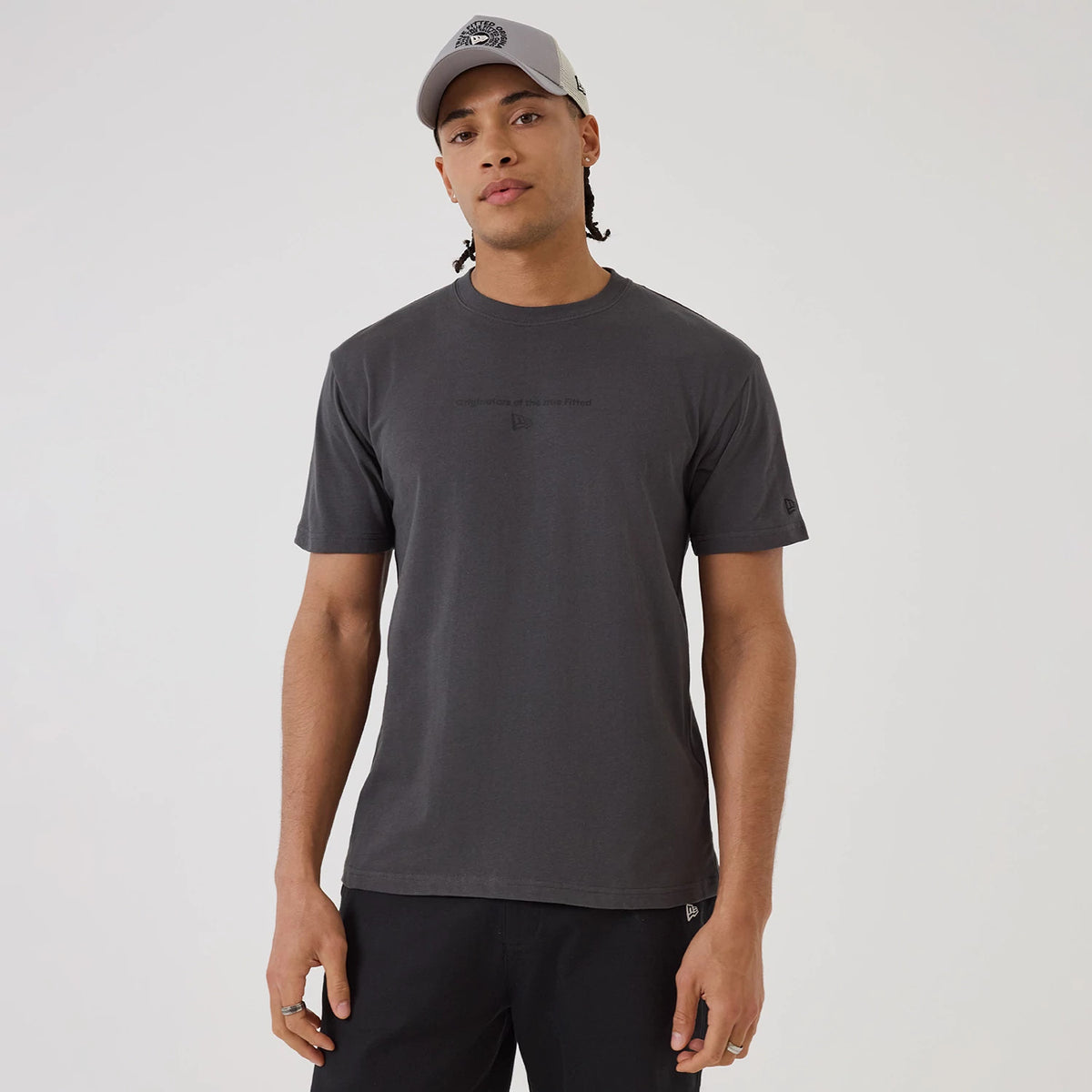New Era Circle Grey T-Shirt, on-model view showing the dark grey short-sleeve design with a small debossed New Era logo on the sleeve, text centred across the chest and a grey cap featuring a raised logo worn by the model.