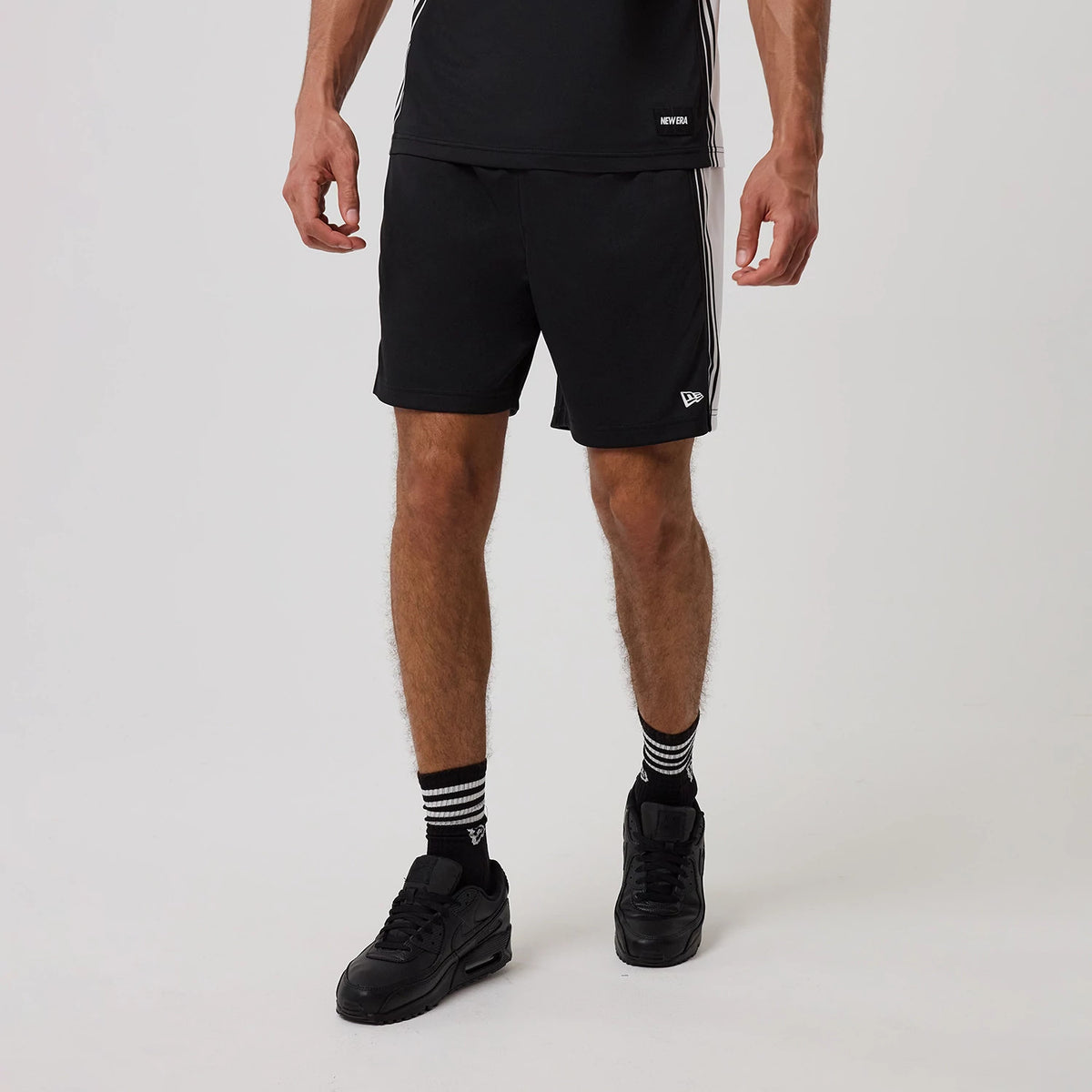 New Era Mesh Black Shorts, on-model view showing the black shorts with a white striped design on the sides, an embroidered New Era logo near the hem on the left leg, worn with matching black trainers and striped socks.