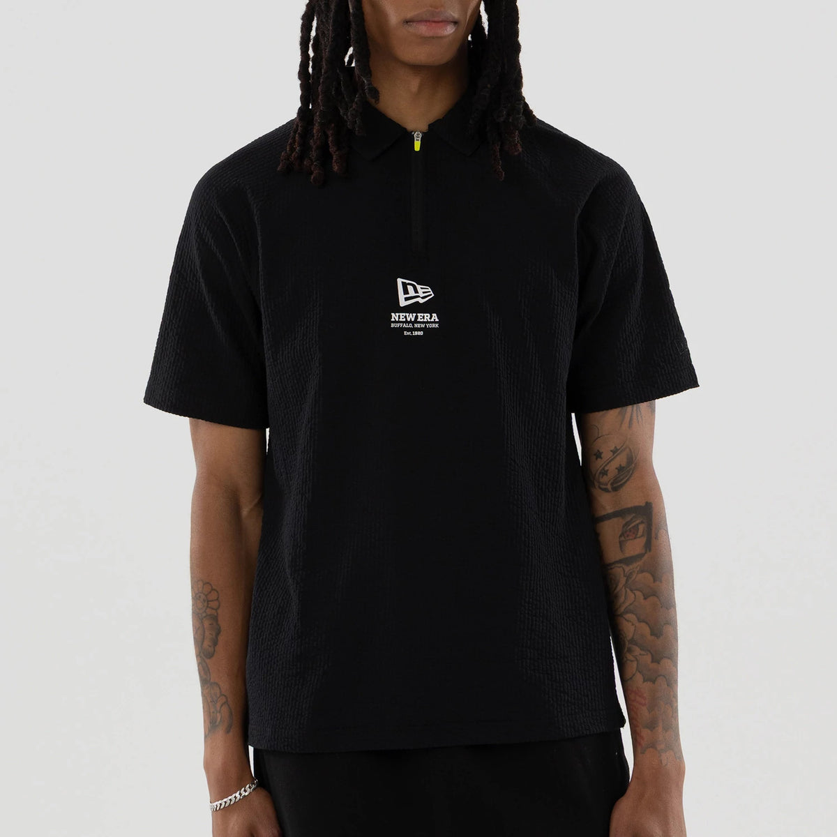 New Era Seersucker Black Polo T-Shirt, front view showing a black polo with short sleeves, a raised New Era logo on the chest below a zip with a contrasting yellow detail, worn by a person with tattoos on the right arm.