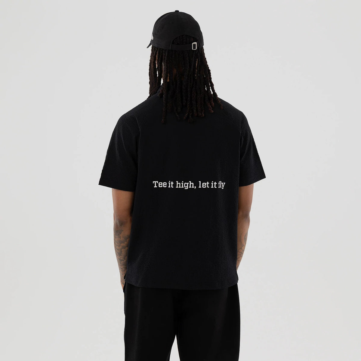 New Era Seersucker Black Polo T-Shirt, rear view showing the phrase "Tee it high, let it fly" in white across the upper back, worn by a model with a black cap featuring a raised logo and an adjustable strap.