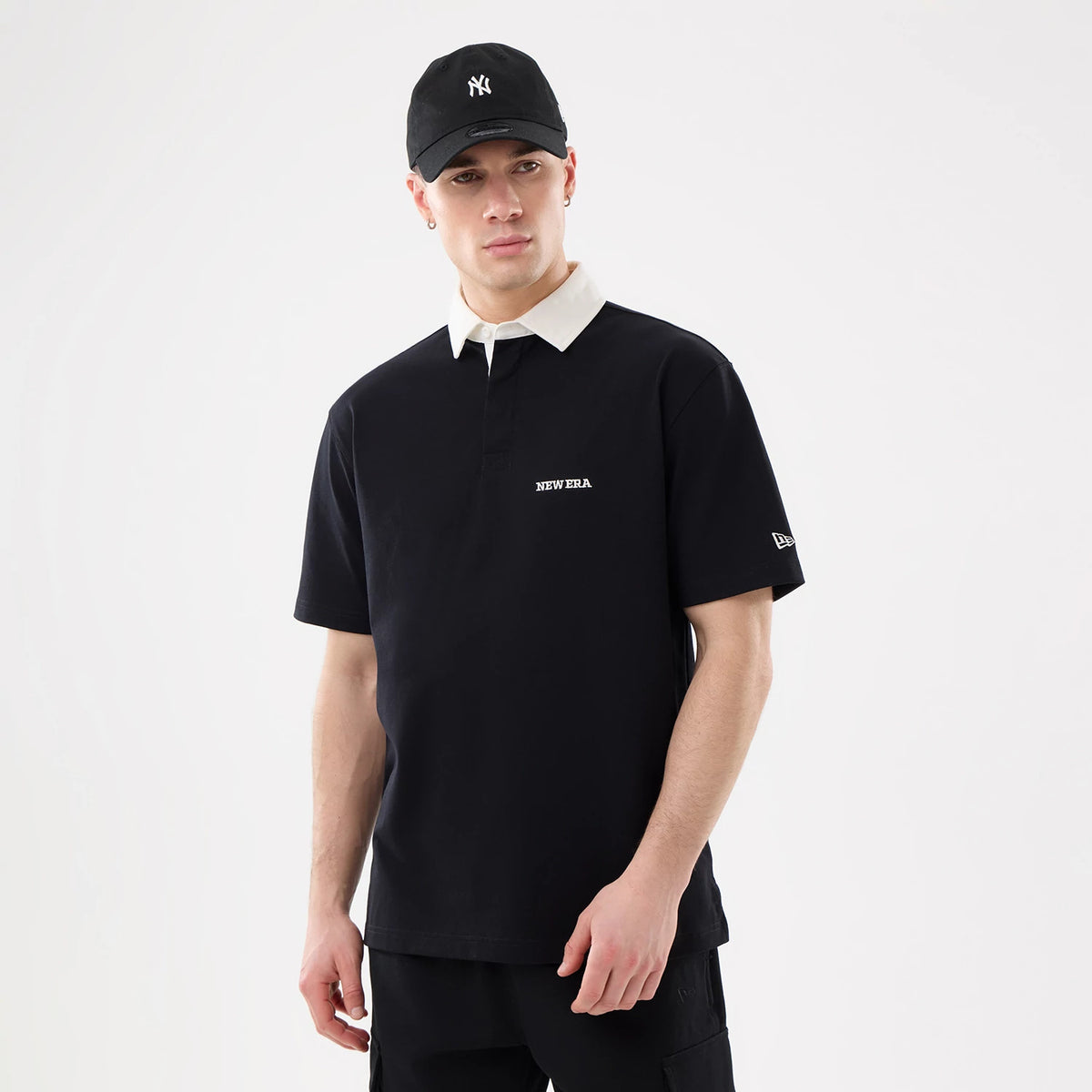 New Era Black Short Sleeve Rugby Shirt, front view on a model wearing a black New York Yankees cap, featuring a white New Era logo on the chest and an embroidered New Era flag logo on the left sleeve.