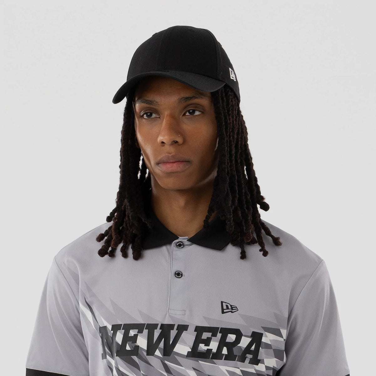 New Era Grid Grey Polo T-Shirt, on-model view showing the grey shirt with a black collar, geometric pattern and New Era text on the chest, a debossed New Era logo on the upper left chest paired with a black cap featuring a raised logo on the side.