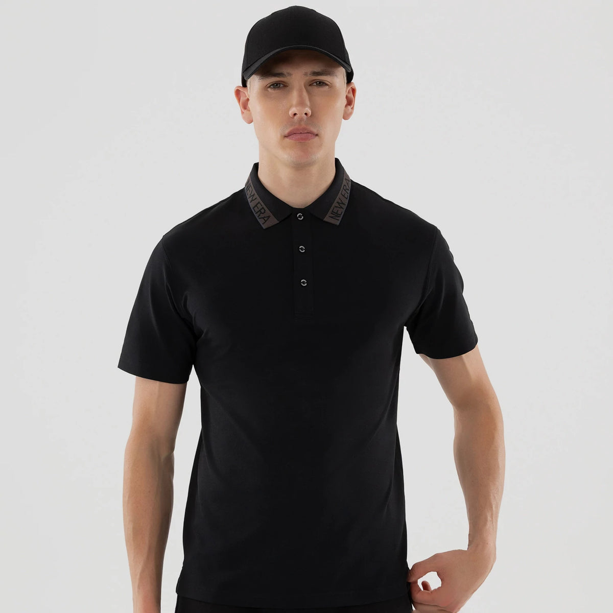 New Era Kanoko Black Polo T-Shirt, front view showing a fitted black polo with a three-button placket, short sleeves, and a New Era logo printed on the collar, worn by a person also wearing a black cap.