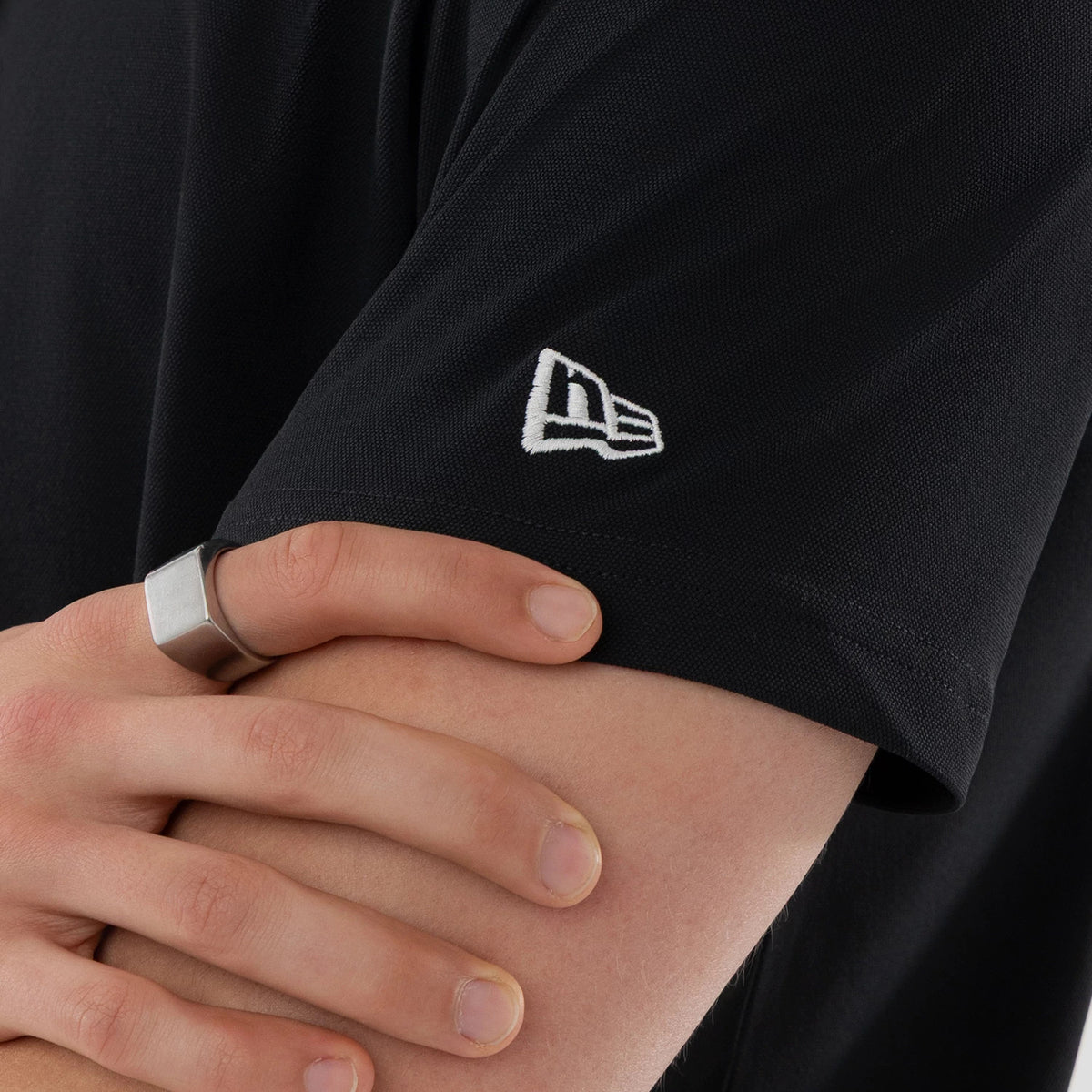 New Era Kanoko Black Polo T-Shirt, close-up view showing the short sleeve with a raised white New Era logo embroidered near the hem, and crossed arms with a silver ring on one hand.