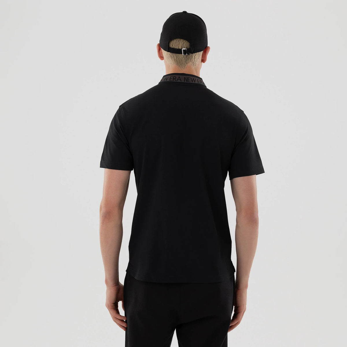 New Era Kanoko Black Polo T-Shirt, rear view showing a black collar with repeated debossed New Era text, short sleeves and a straight hem, worn by a person also wearing a black cap with an adjustable strap.