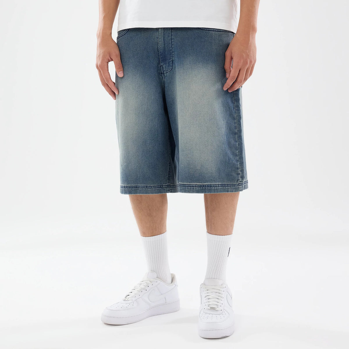 New Era Washed Blue Bermuda Shorts, front view showing a faded blue design with knee-length hem, subtle stitching details on the hems and pockets, worn by a model with white sneakers and socks.
