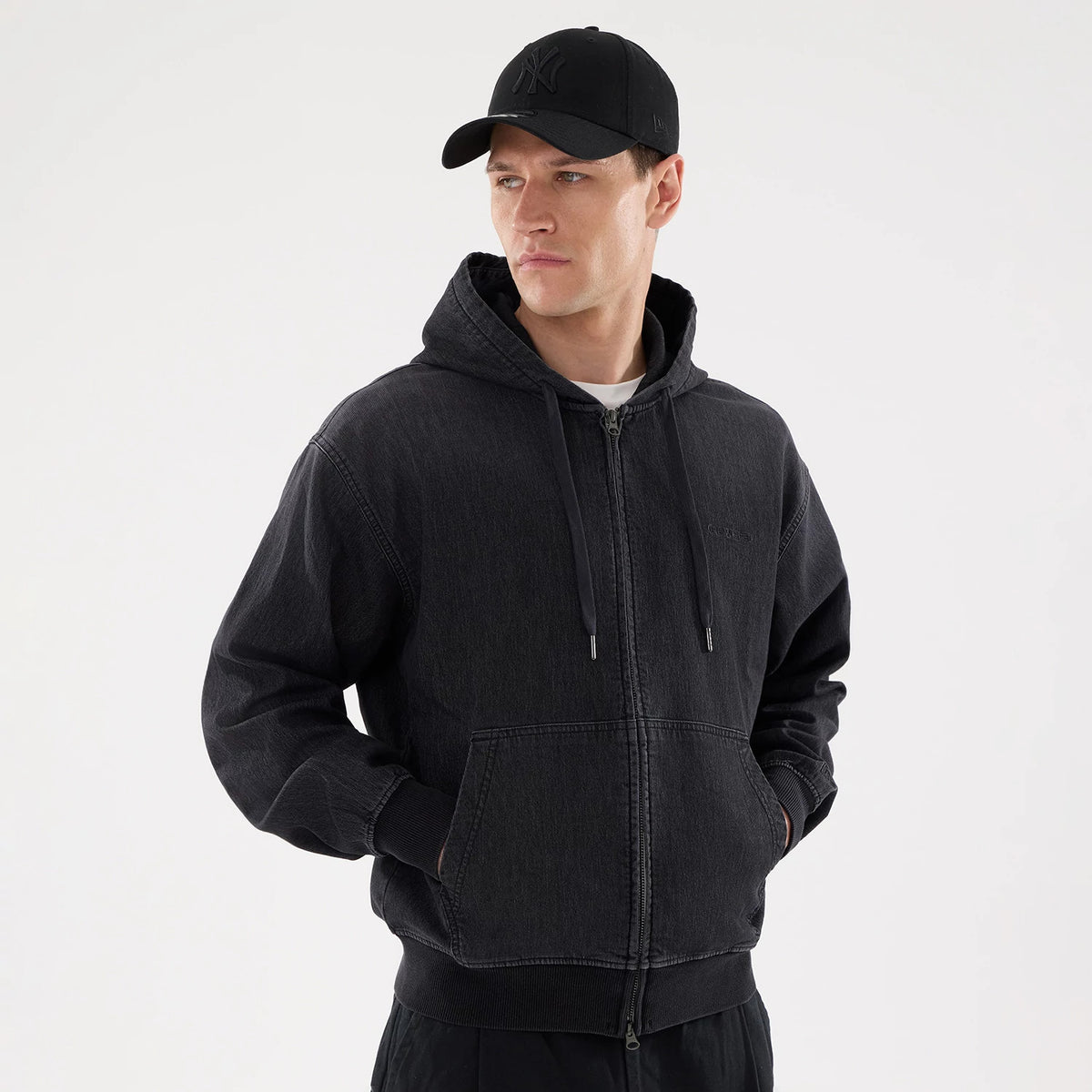 New Era Washed Black Full Zip Hoodie, on-model view showing a front zip closure, adjustable drawstring hood, ribbed cuffs, and a kangaroo pocket, paired with a black cap featuring a raised logo on the front.
