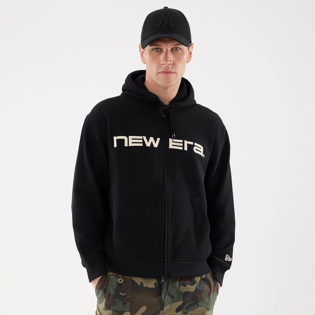 New Era Waffle Black Full Zip Hoodie, front view showing large white New Era text logo across the chest, attached hood with drawstrings, full-length zip closure, and black embroidered New York logo on a black cap worn by the model.