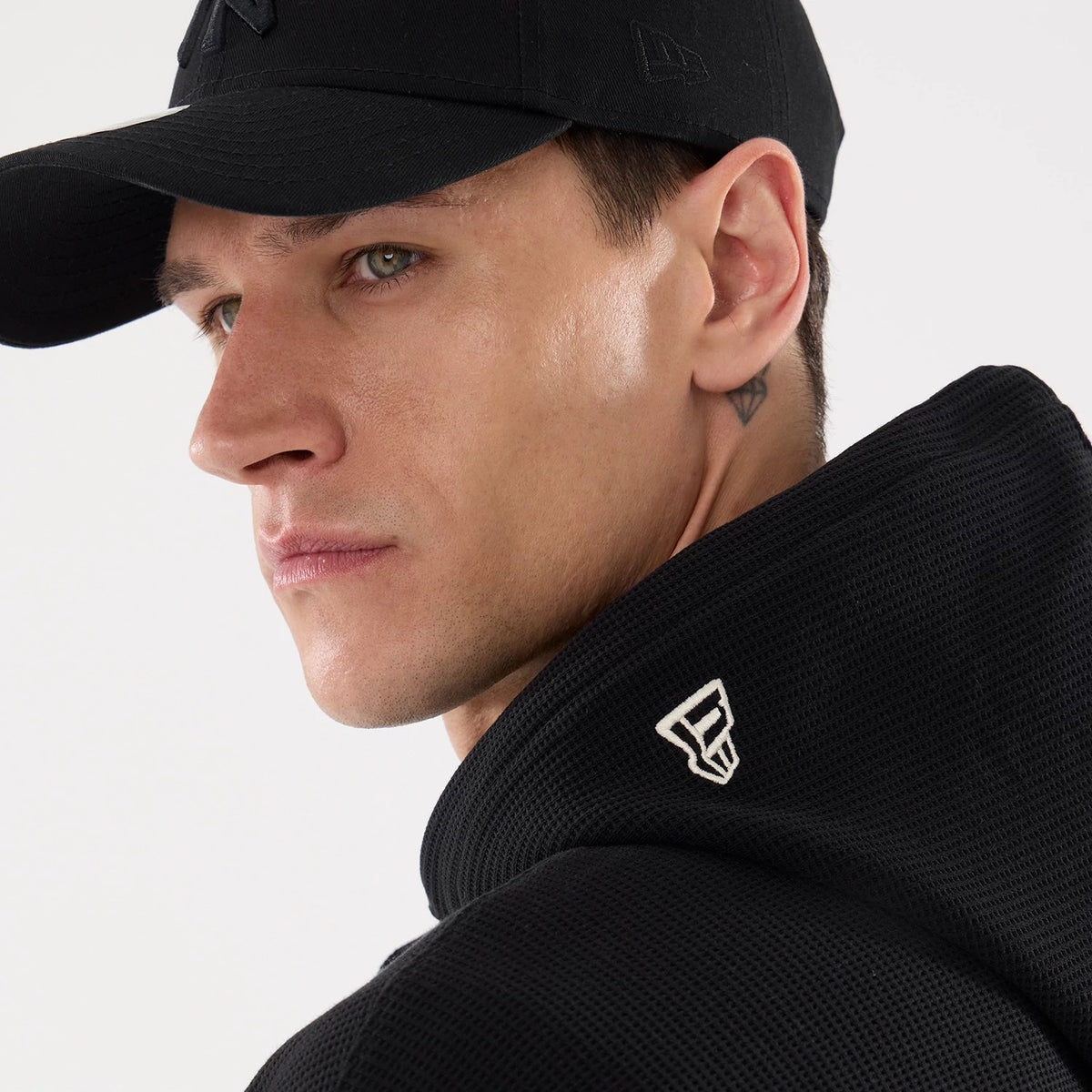 New Era Waffle Black Full Zip Hoodie, on-model view showing the textured black fabric with a white debossed New Era logo on the raised collar, paired with a black cap featuring a debossed logo on the side.