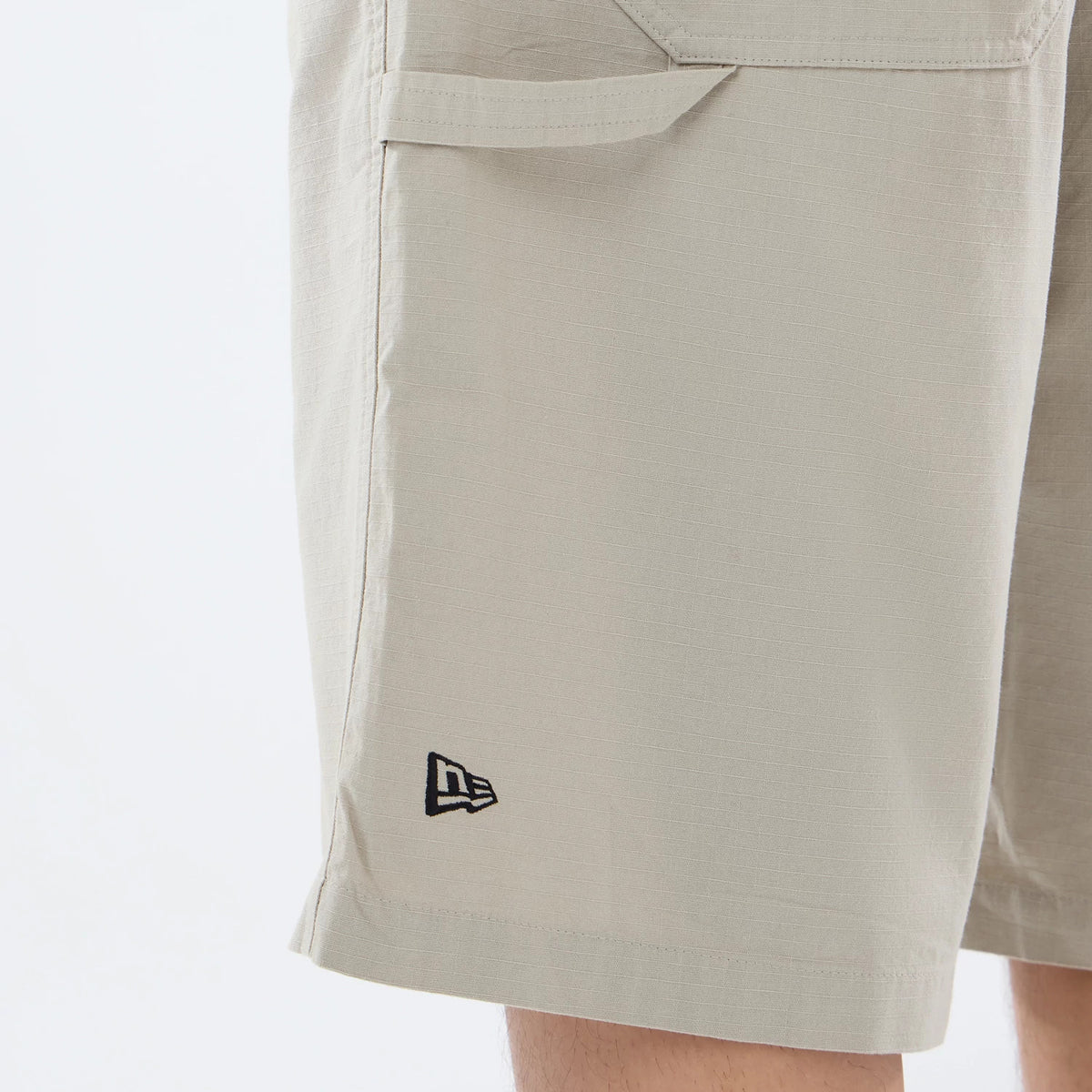 New Era Canvas Beige Bermuda Shorts, side view showing the light beige fabric with a debossed black New Era logo near the hem and a large pocket with flap detail at the upper side seam.