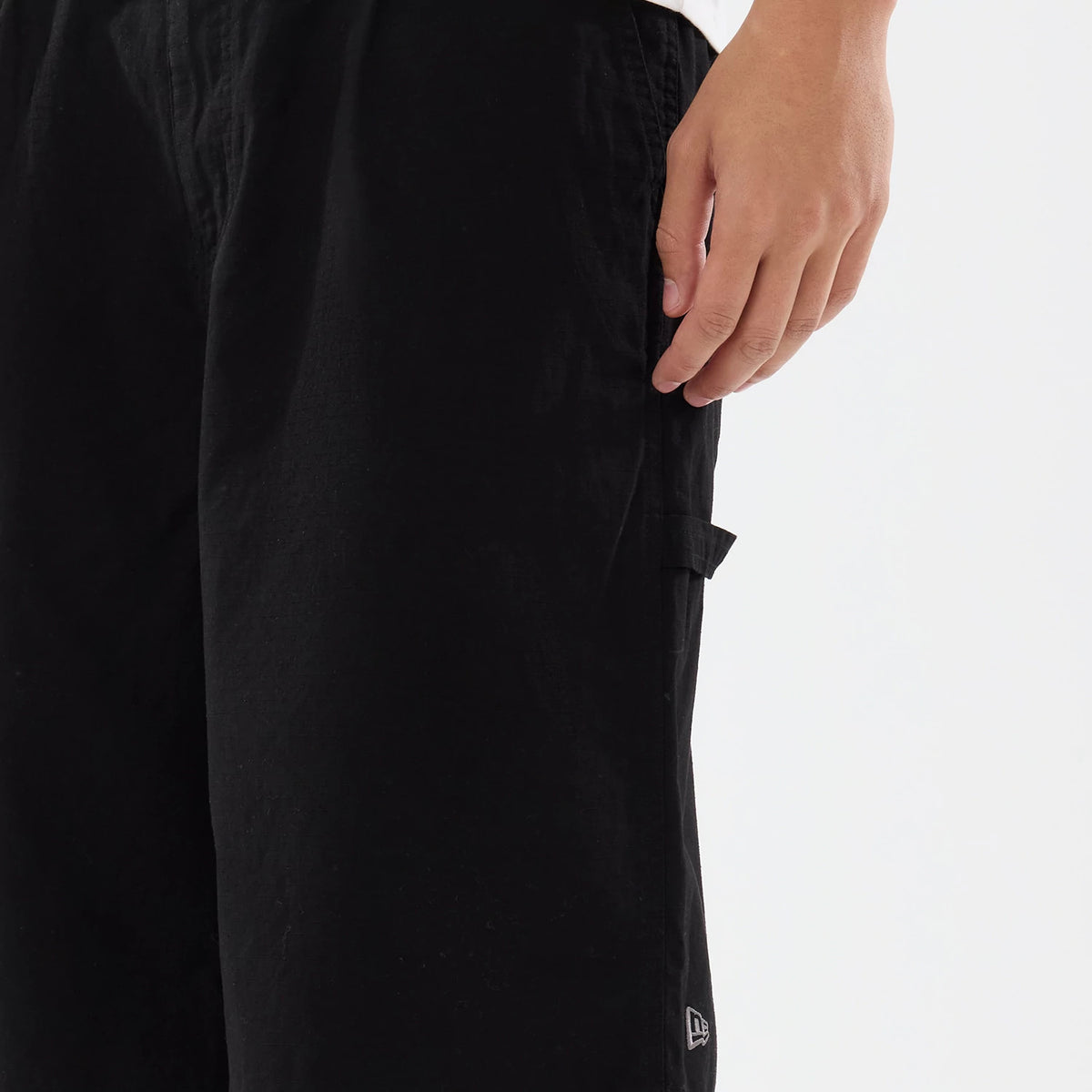 New Era Canvas Black Bermuda Shorts, side view showing a close-up of the lower part with a debossed New Era logo near the hem on the left leg and a hand resting lightly on the right side above a flap pocket.