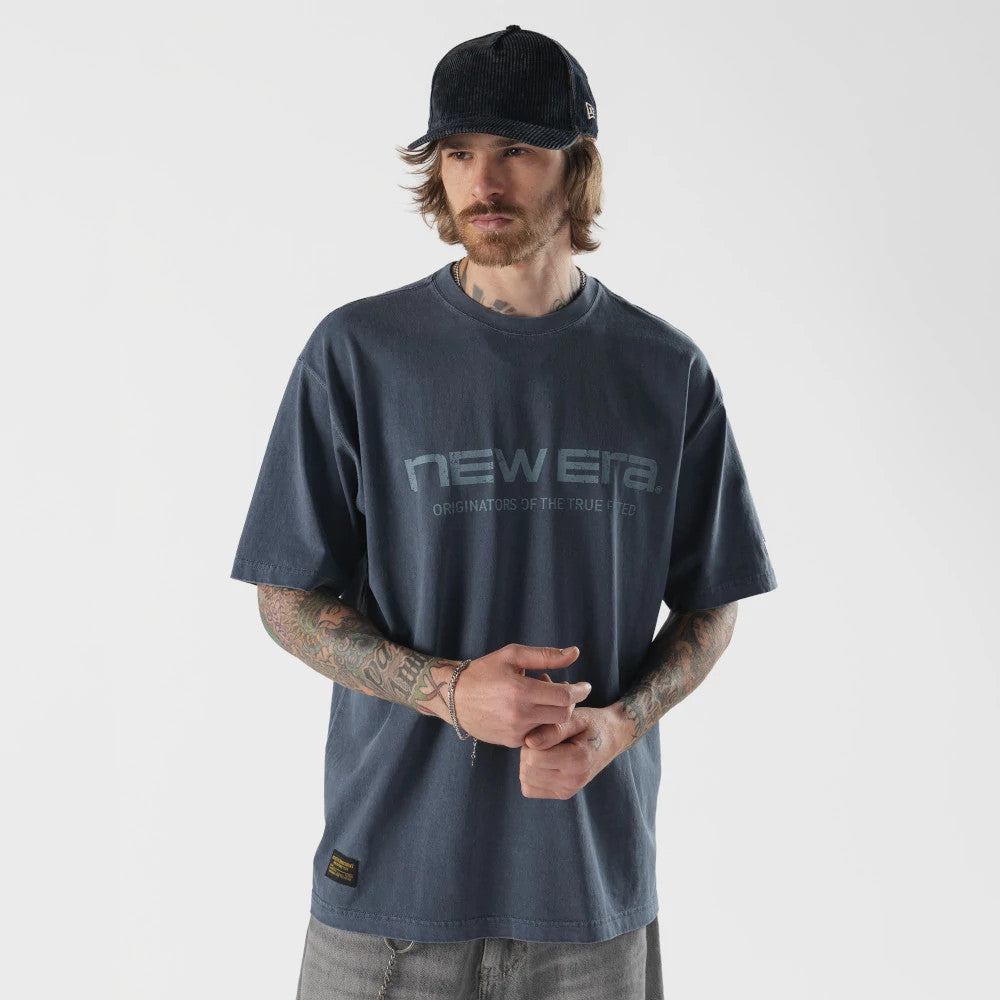 New Era Originators Navy T-Shirt, on-model view showing the navy shirt with a large printed New Era logo across the chest, short sleeves, and a small woven patch near the hem, paired with a navy cap.