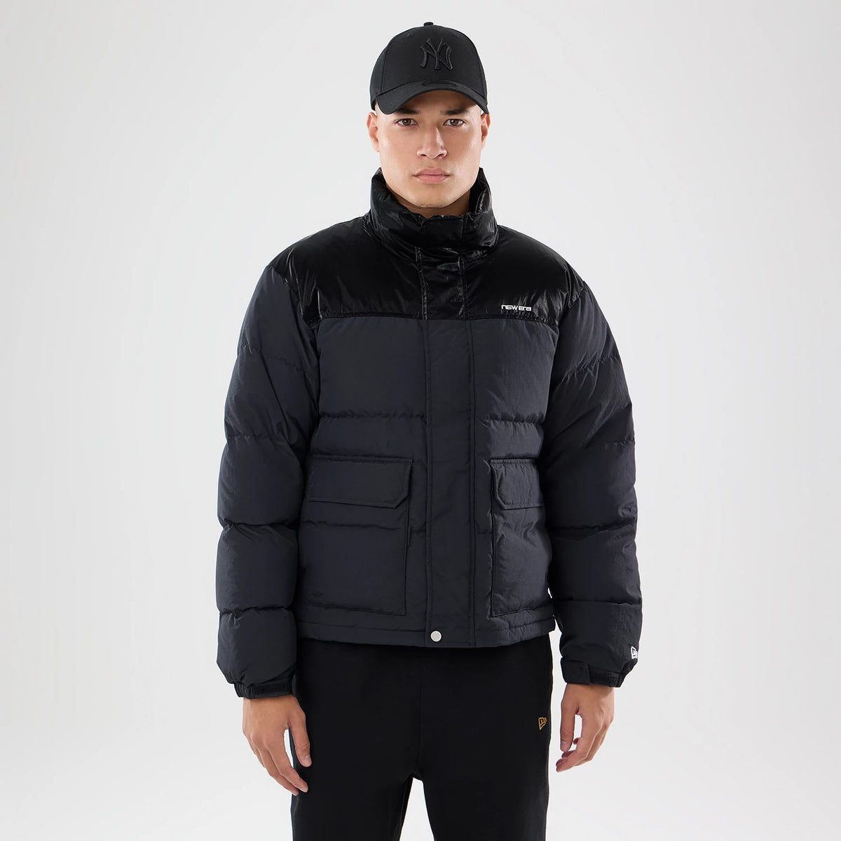 New Era Down Black Jacket, front view showing the puffy black jacket with a high collar, two flap pockets on the front, a small embroidered New Era logo on the left chest, and the jacket worn by a person wearing a black cap.