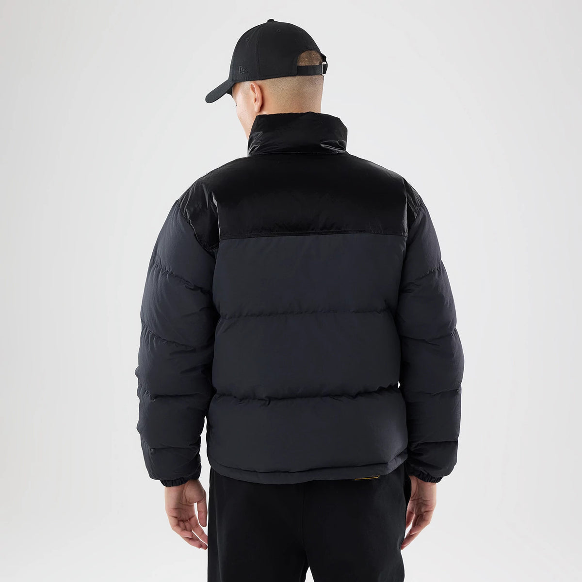 New Era Down Black Jacket, rear view showing a quilted puffer design with a high collar, the black jacket worn by a model in black trousers, complemented by a black cap with a raised New Era logo on the side.