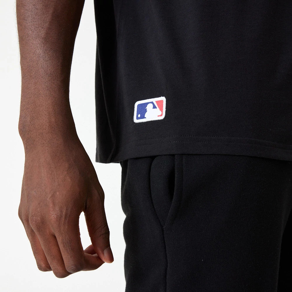 New York Yankees MLB Essential Black Oversized T-Shirt, side view showing a black hem with a colourful embroidered MLB logo near the bottom, paired with matching black trousers on the model.