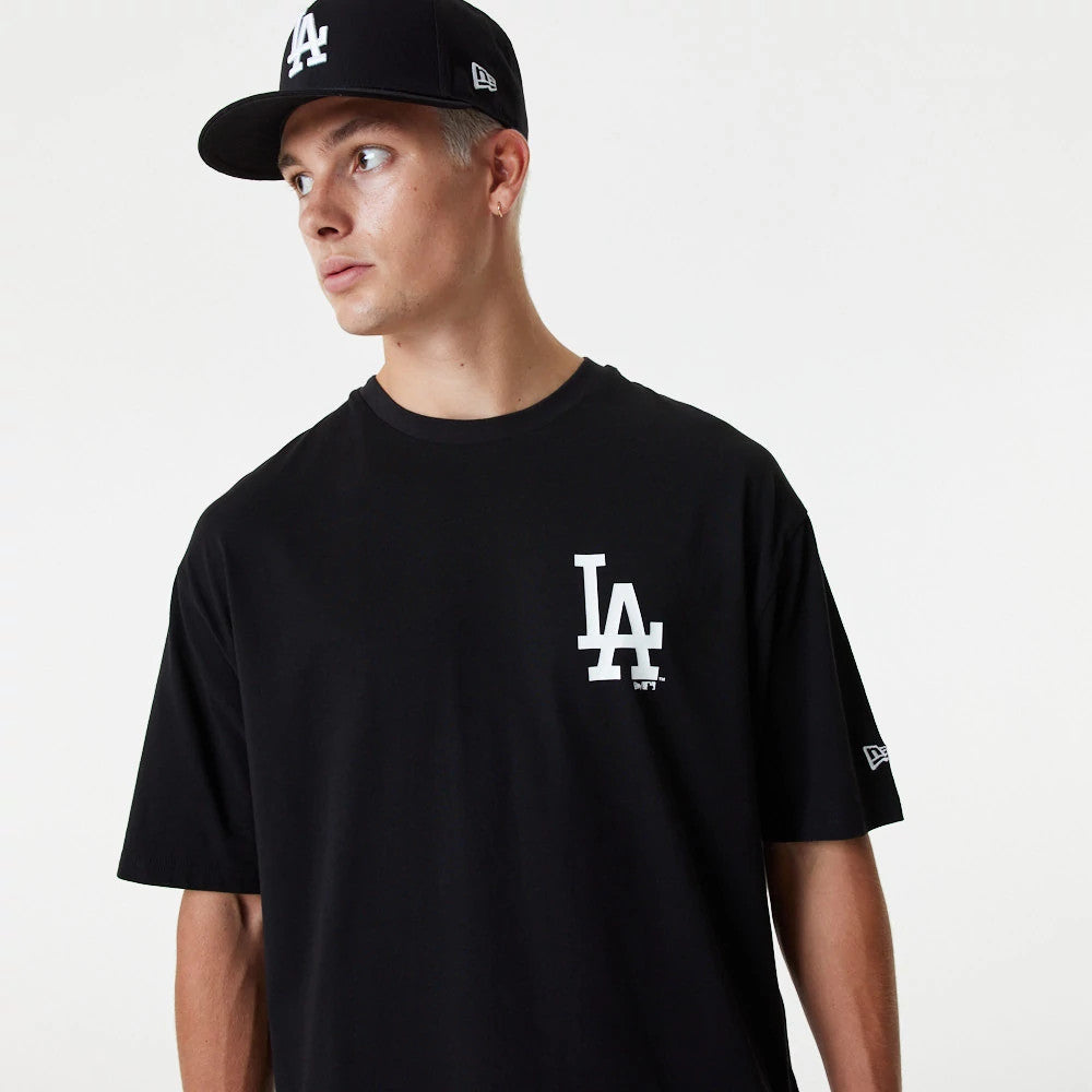 LA Dodgers MLB Essential Black Oversized T-Shirt, on-model view showing a large white LA logo on the left chest, worn by a person paired with a black cap featuring a raised LA logo on the front and a New Era logo on the side.