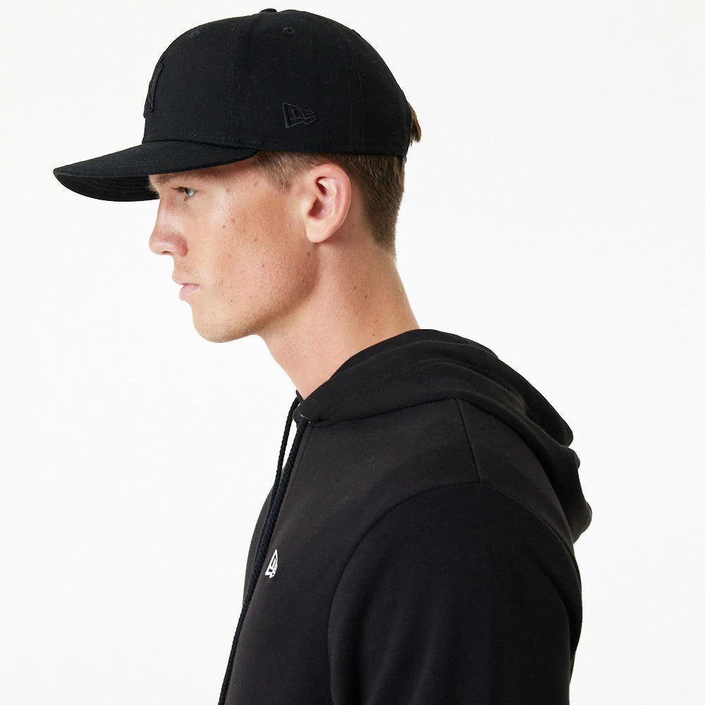 New Era Essential Black Pullover Hoodie, on-model side view showing the black hoodie with a small embroidered logo on the chest and a black cap featuring a debossed New Era logo on the side panel.