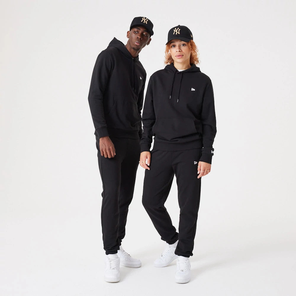 New Era Essential Black Pullover Hoodie, on-model view featuring two individuals wearing black hoodies with raised New Era logos on the chest near the shoulder, both paired with black trousers and black caps showing the raised New York Yankees logo.