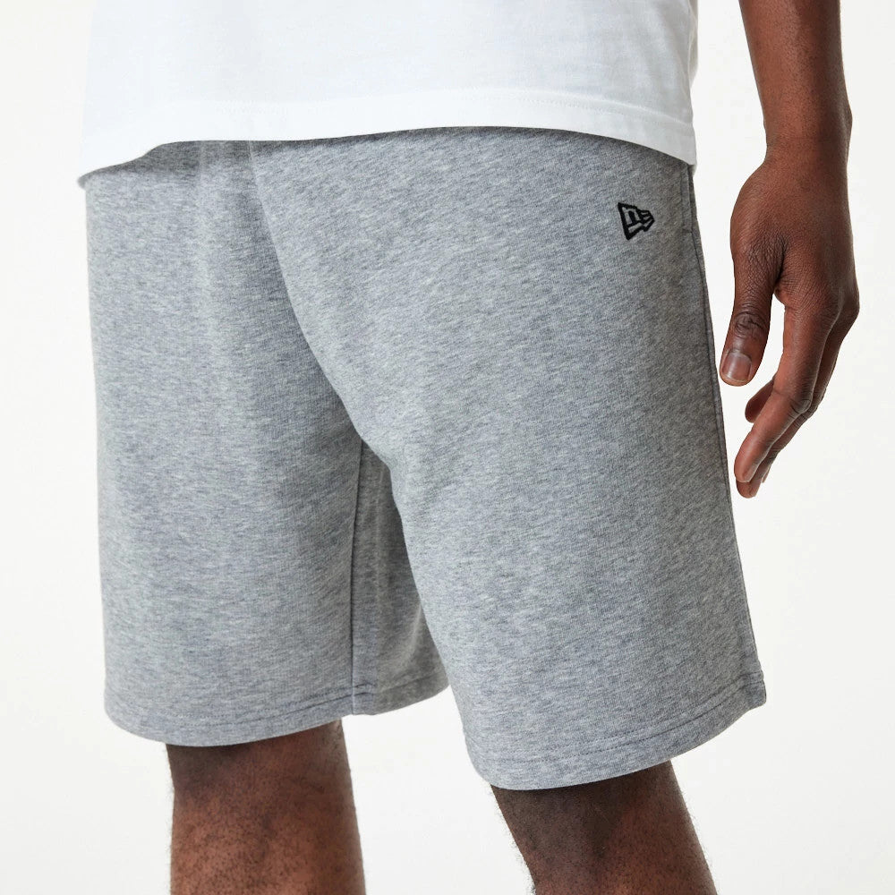 New Era Essential Grey Shorts, side view showing the light grey fabric with a small black raised New Era logo on the upper left thigh and a relaxed fit extending to just above the knee.