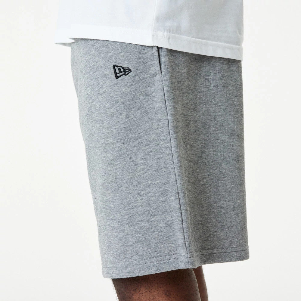 New Era Essential Grey Shorts, side view showing light grey fabric with a raised New Era logo in black near the pocket seam, the hem falling to knee length against a neutral backdrop.