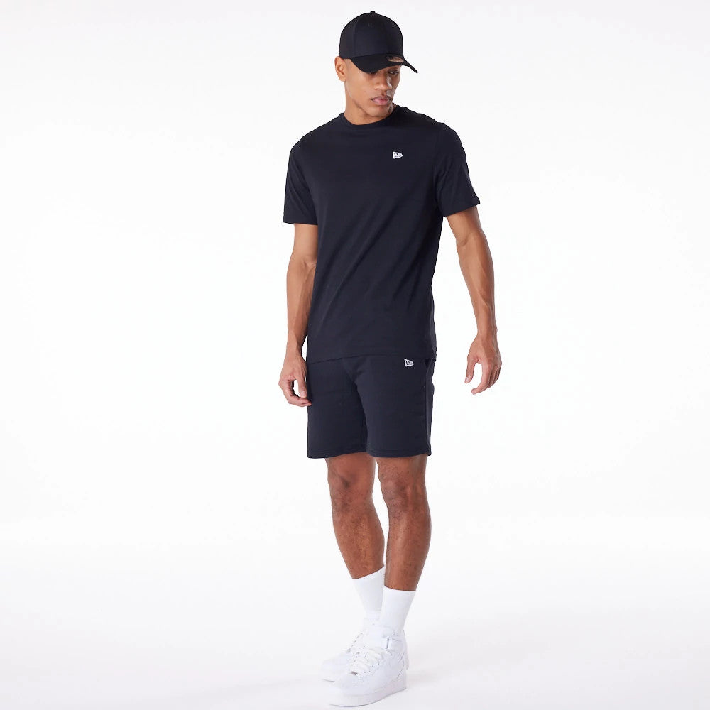 New Era Essential Black Logo Shorts, on-model view showing the black shorts with a raised logo on the left leg, paired with a matching black t-shirt featuring the same logo on the chest, and a black cap worn by the model.