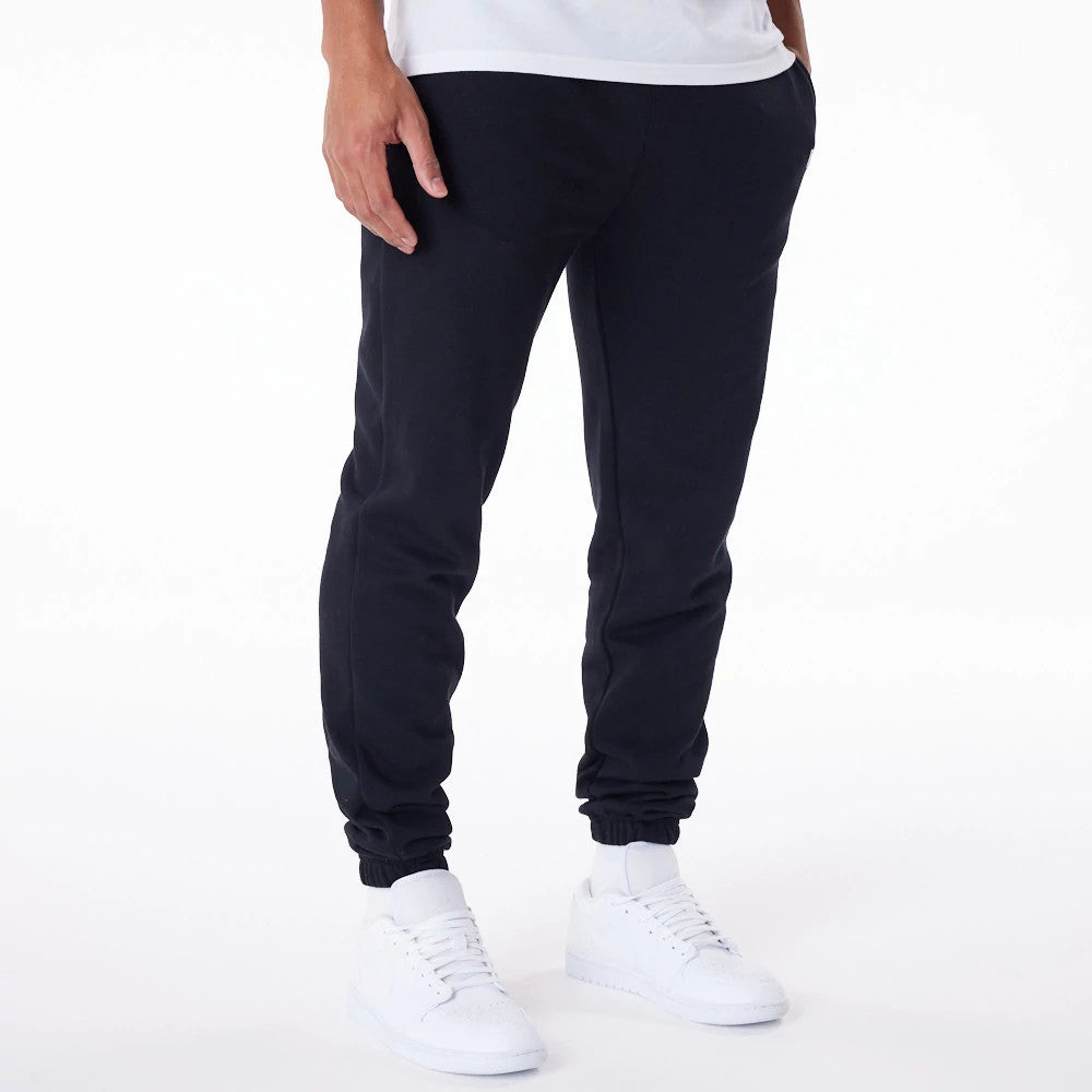 New Era Essential Black Joggers, front view on a model wearing a white T-shirt and white trainers, showing a relaxed fit with ribbed ankle cuffs and side pockets on soft black fabric.