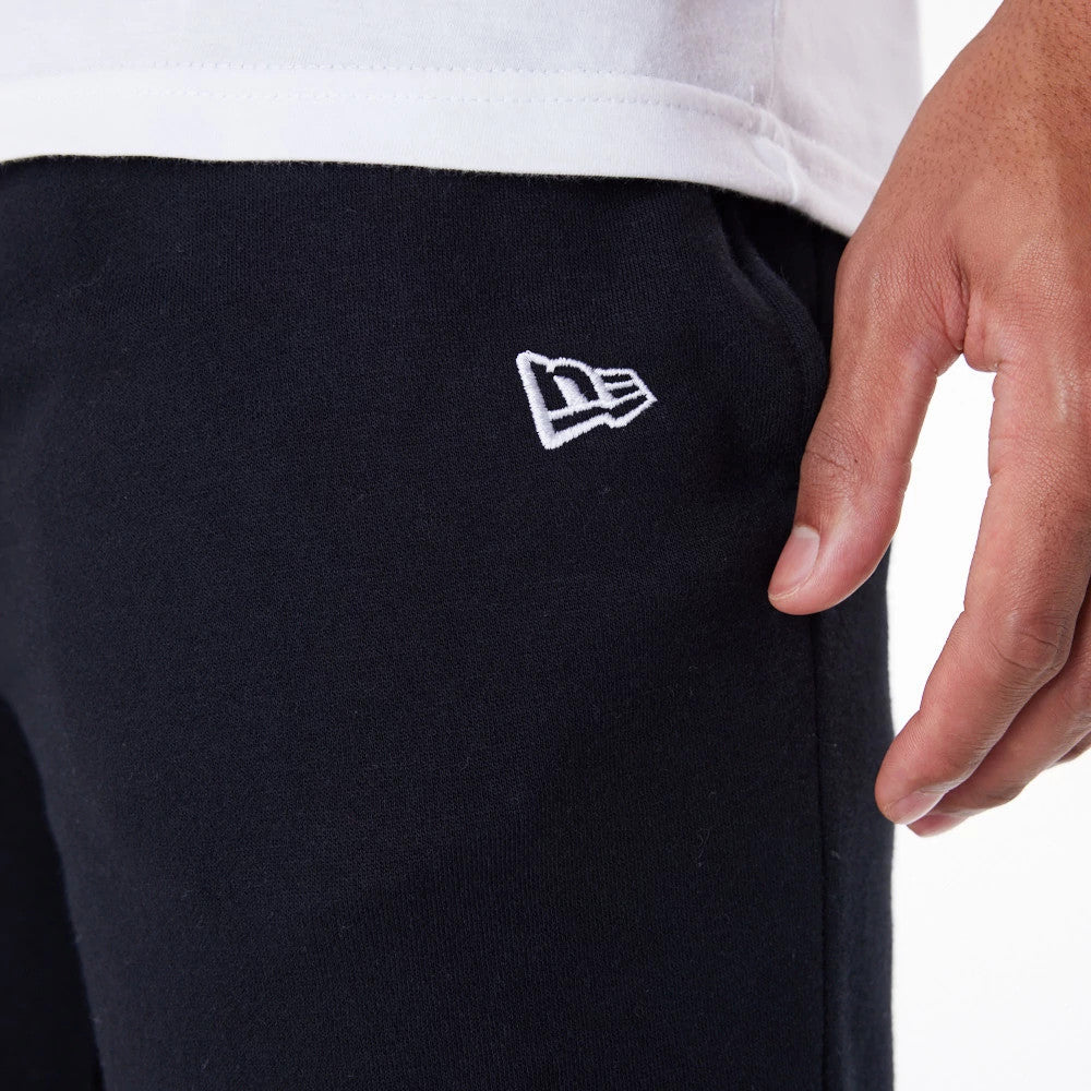 New Era Essential Black Joggers, close-up view on a model showing the white New Era flag logo embroidered on the left thigh, paired with a white shirt and detail of a hand by the pocket.